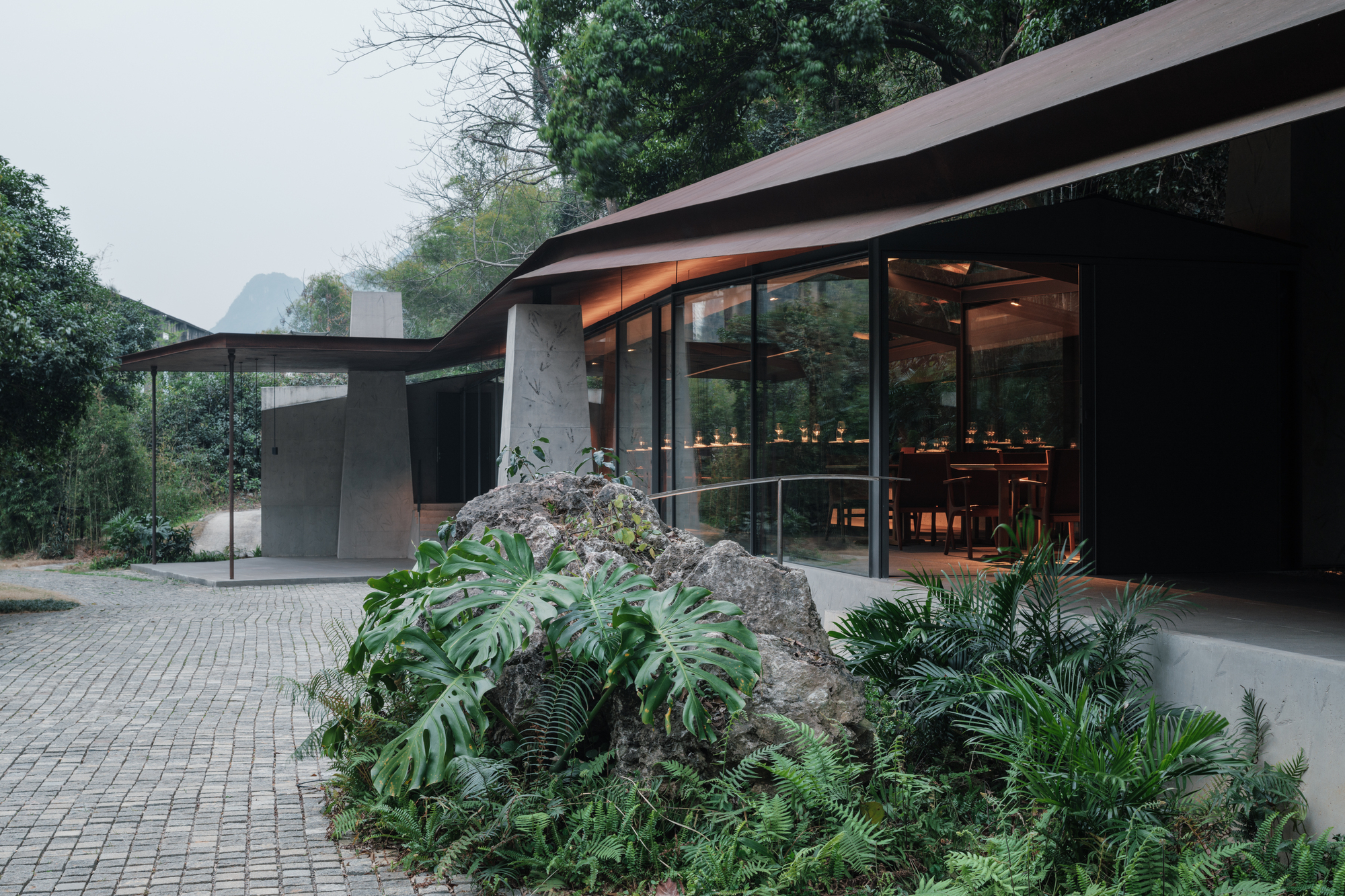 Glazed dining pavilion under a folded dark roof, with a mossy boulder and ferns in the foreground and cobbled paving beside it