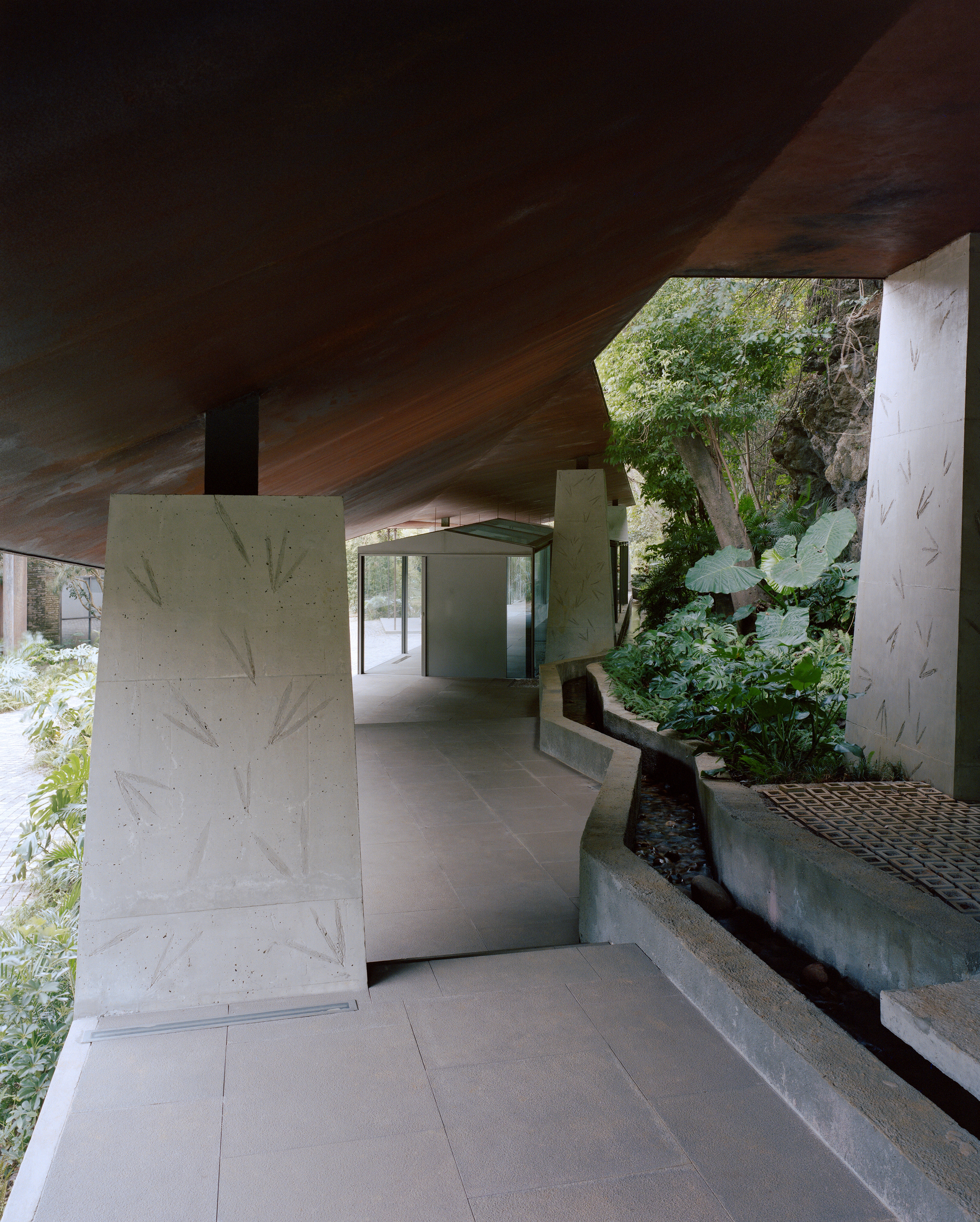 Covered walkway beneath a sloping rusted-steel roof, with leaf-imprinted concrete piers and a narrow water channel beside the planted bank