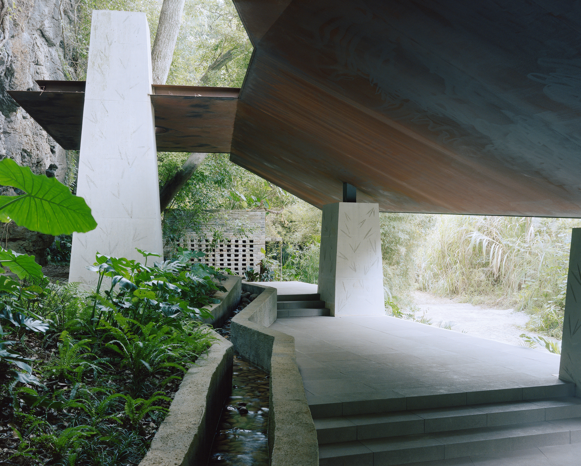 Stepped concrete walkway running beside a shallow stream channel, with tall leaf-patterned piers and ferns under a cantilevered timber-toned roof