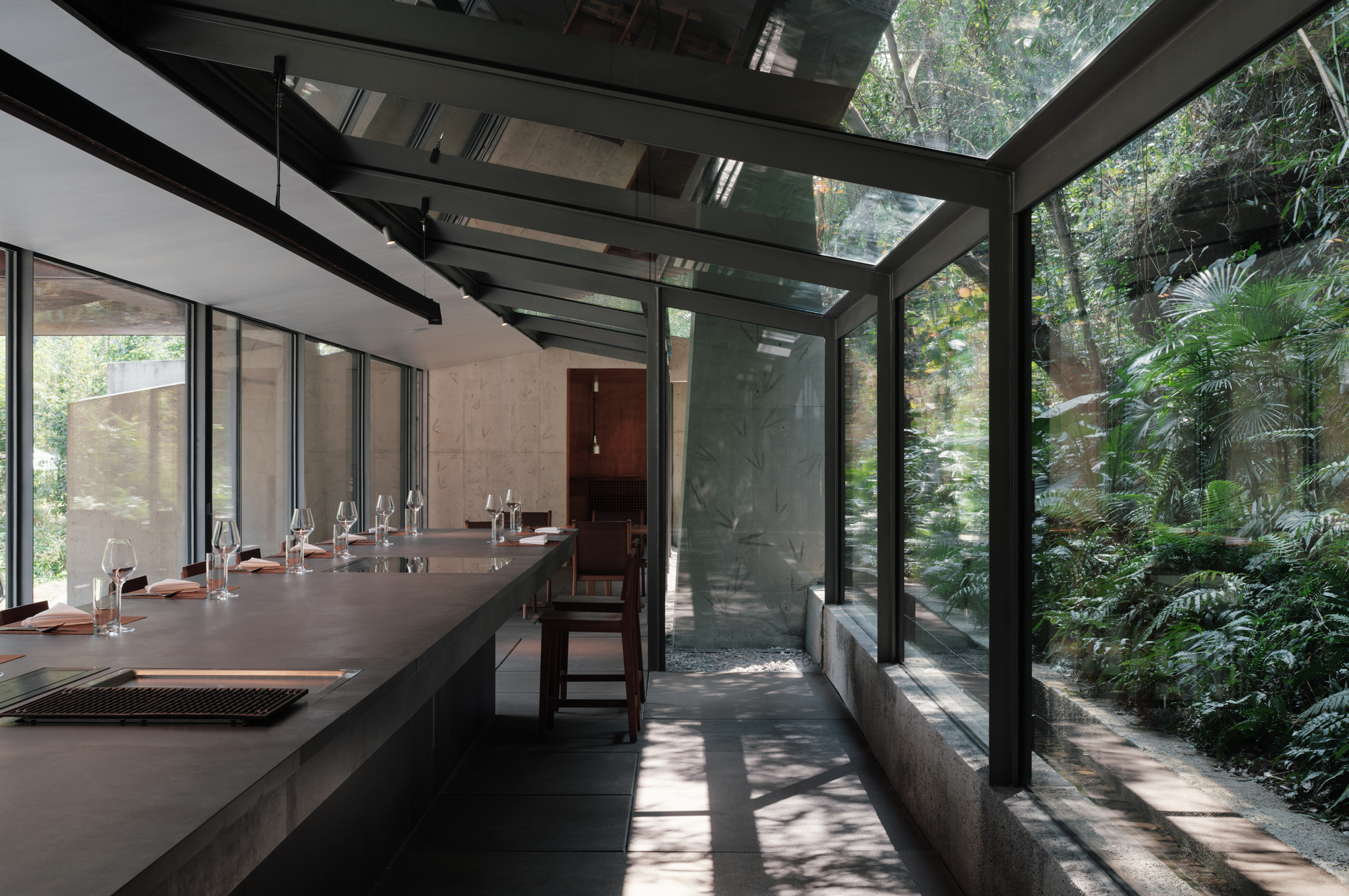 Long dark counter with place settings beside a glass-roofed corridor, dappled sunlight on the floor and dense palms outside