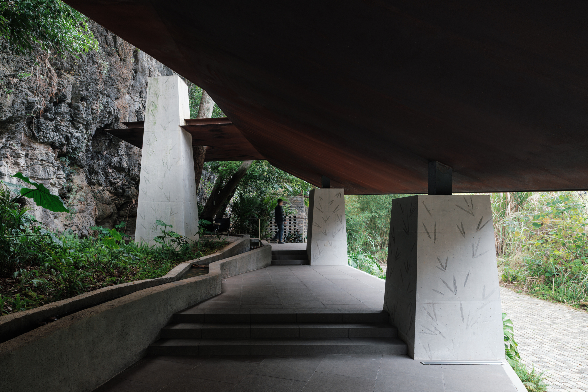 Concrete steps and a low channel wall leading past tapered piers toward a limestone cliff, with a lone figure walking in the distance