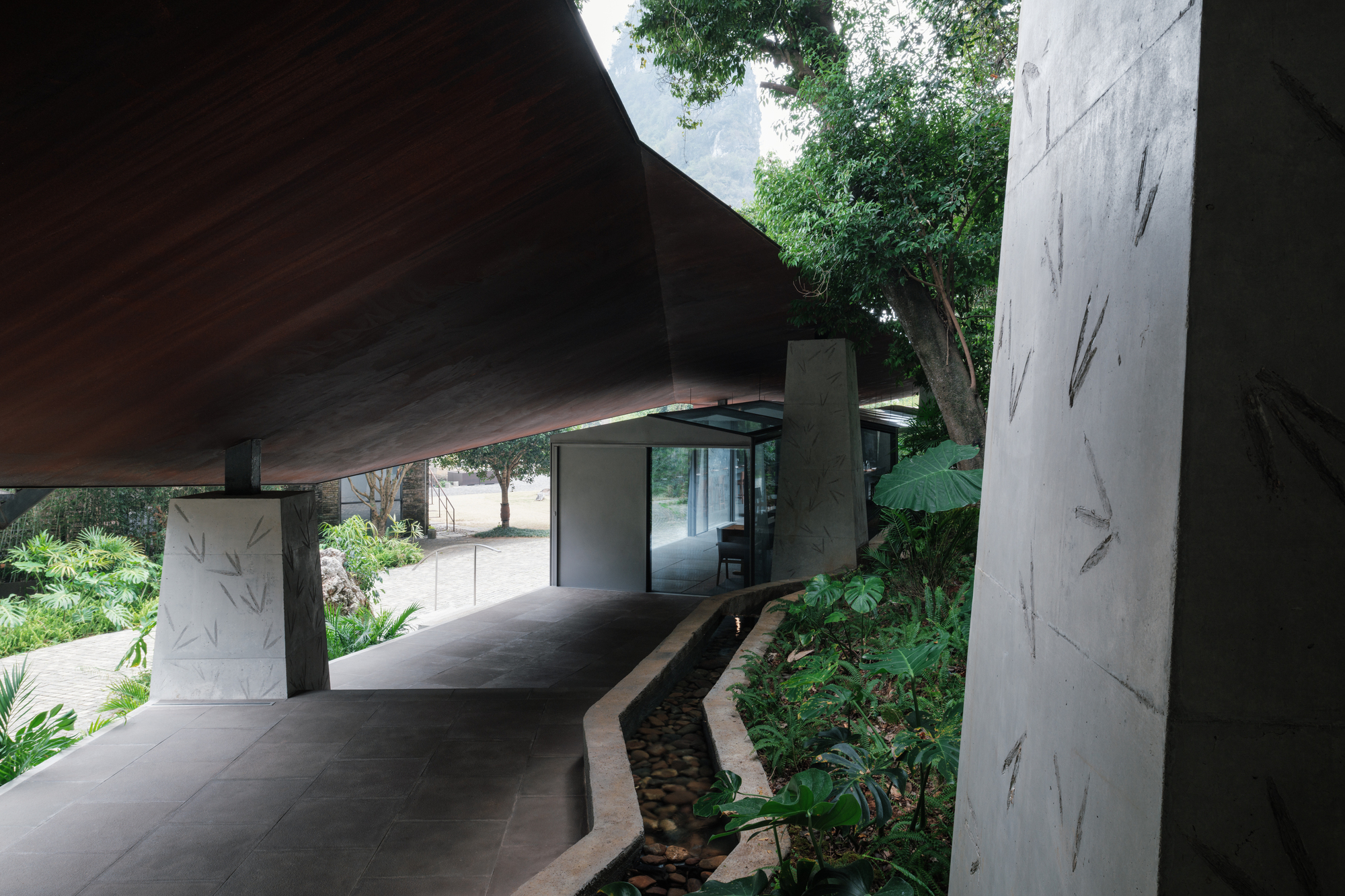 Overhanging roof above a paved passage with a pebble-lined water channel, a large tree and a glass pavilion ahead