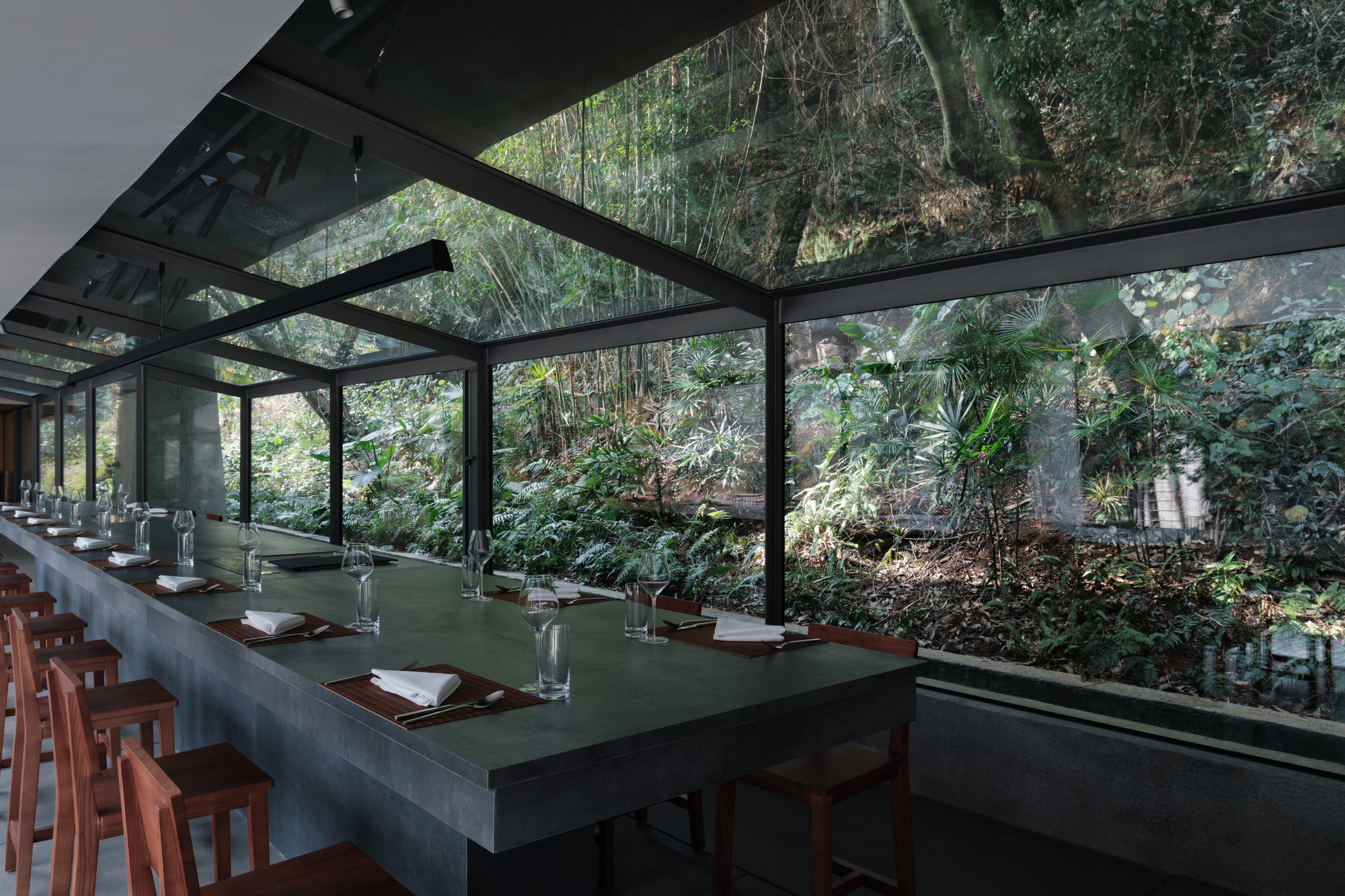Long dark stone dining counter beside a glass wall facing a planted slope of ferns and bamboo