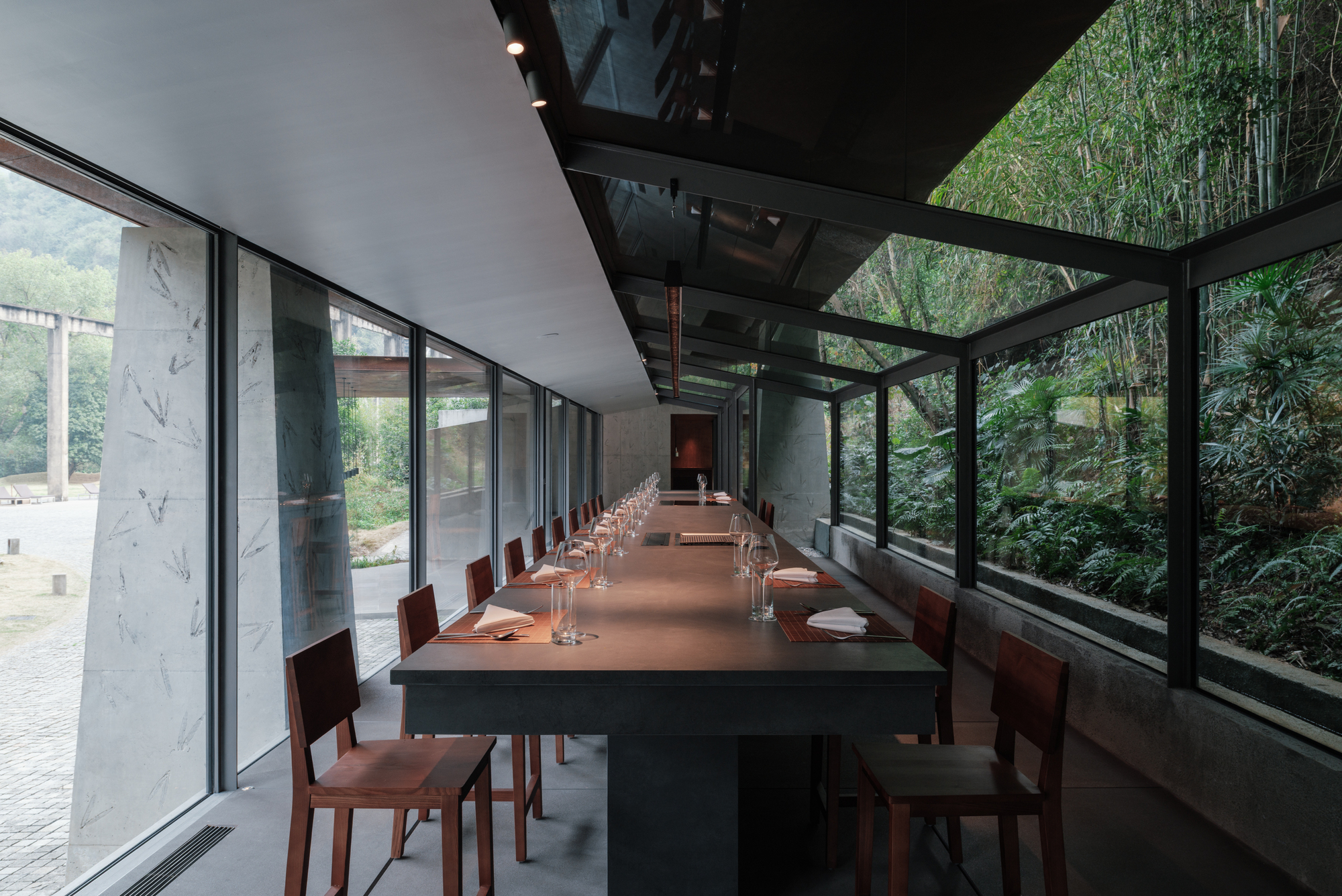 Narrow dining room with a long counter, timber chairs and a sloped glass roof against a bamboo hillside