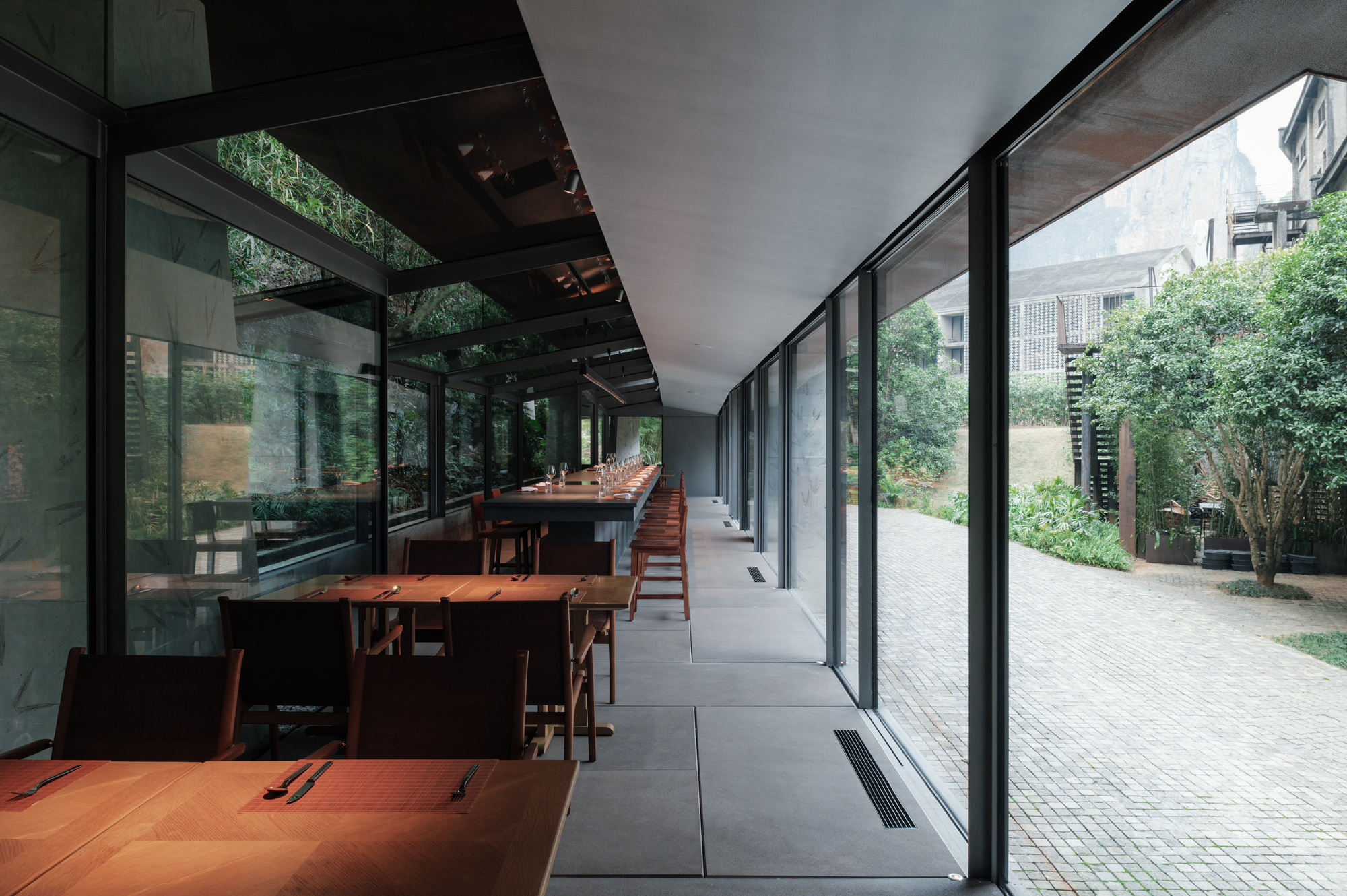 Side aisle with timber tables and chairs running along floor-to-ceiling glass that opens onto a brick-paved courtyard