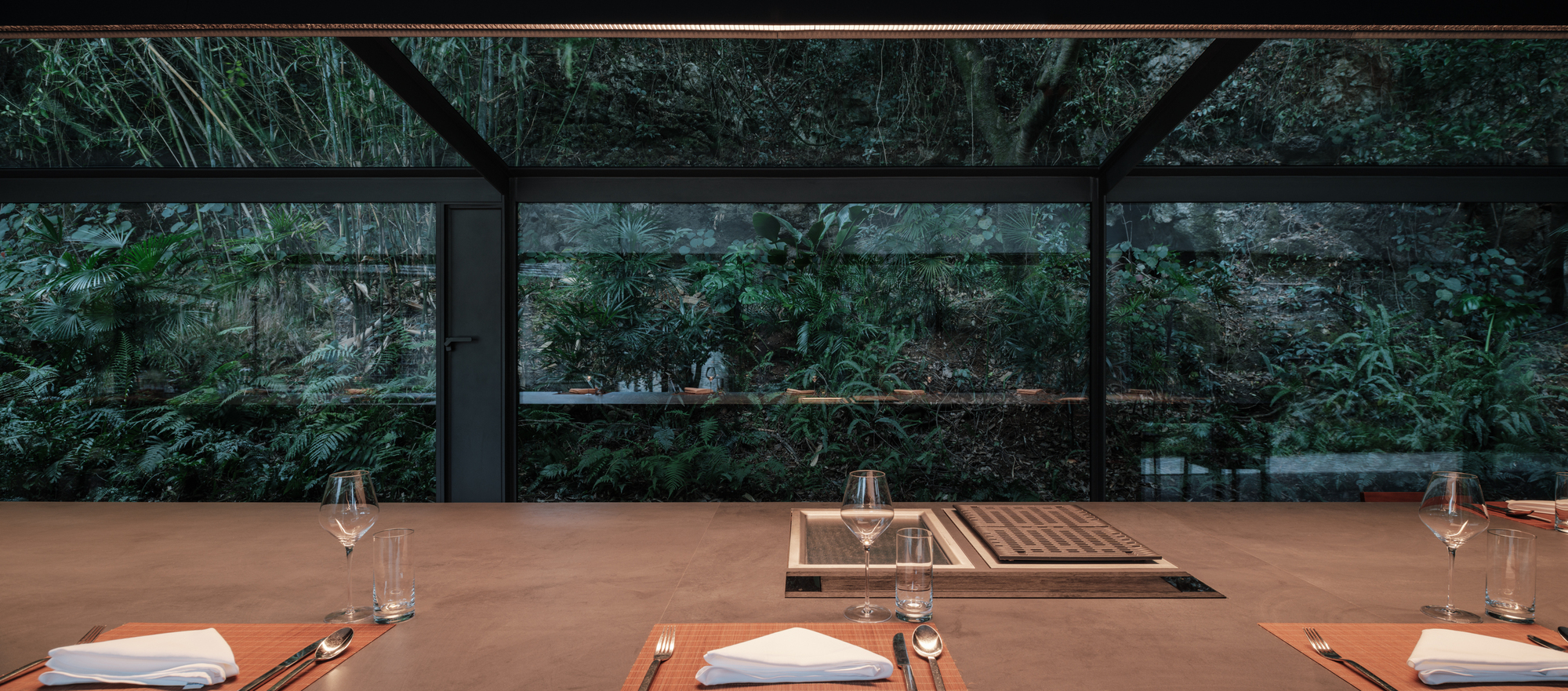 Table set with napkins, cutlery and glasses beside a recessed grill, facing floor-to-ceiling glass and dense green foliage