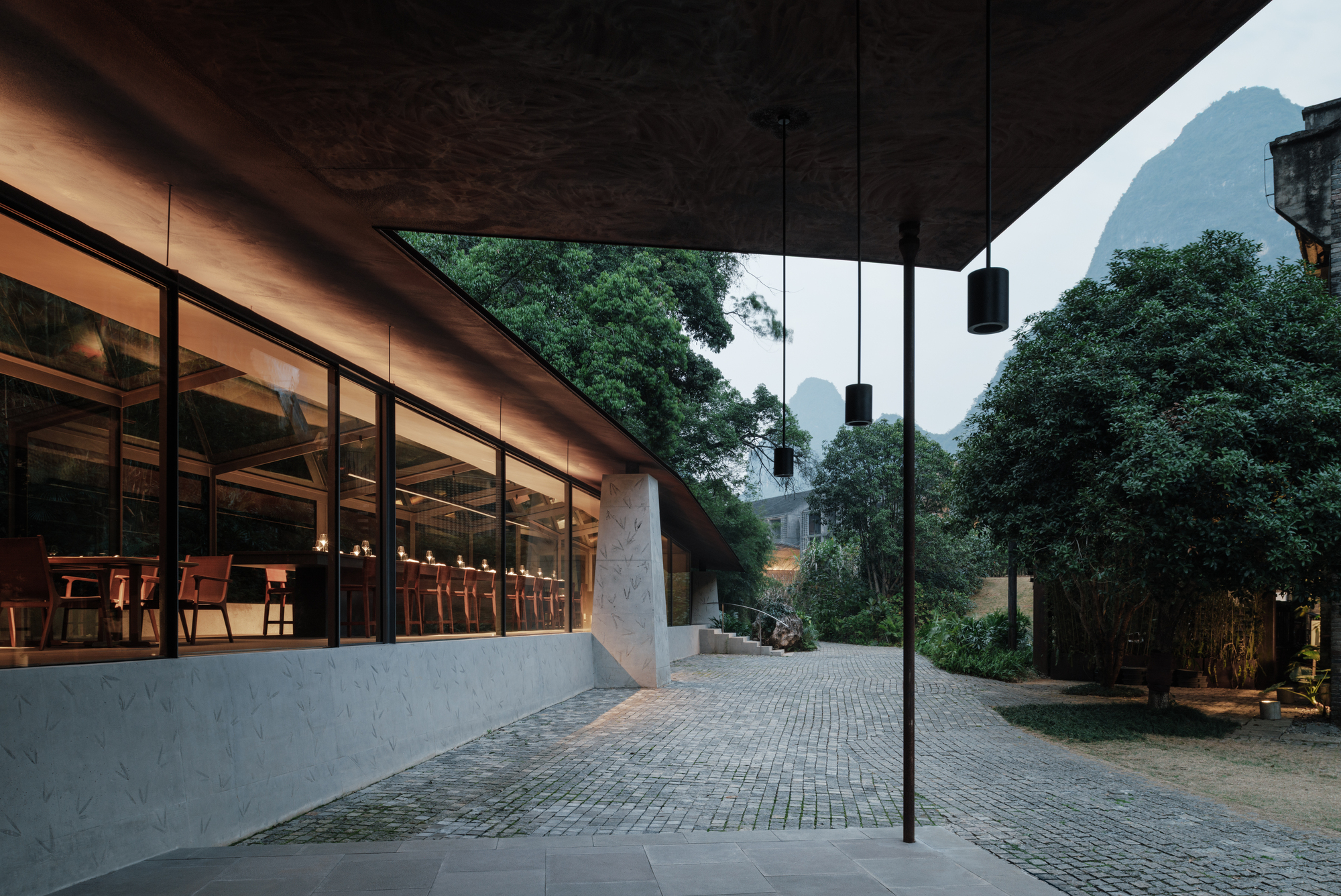 Covered edge of the glazed dining hall with a pale stone wall, cobbled courtyard and hazy limestone peaks behind trees at dusk