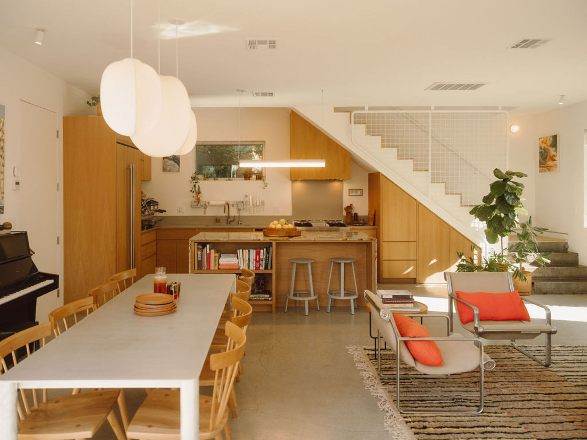 Open living area with a long white dining table, spindle-back chairs and a timber kitchen island beneath a white staircase in afternoon sun