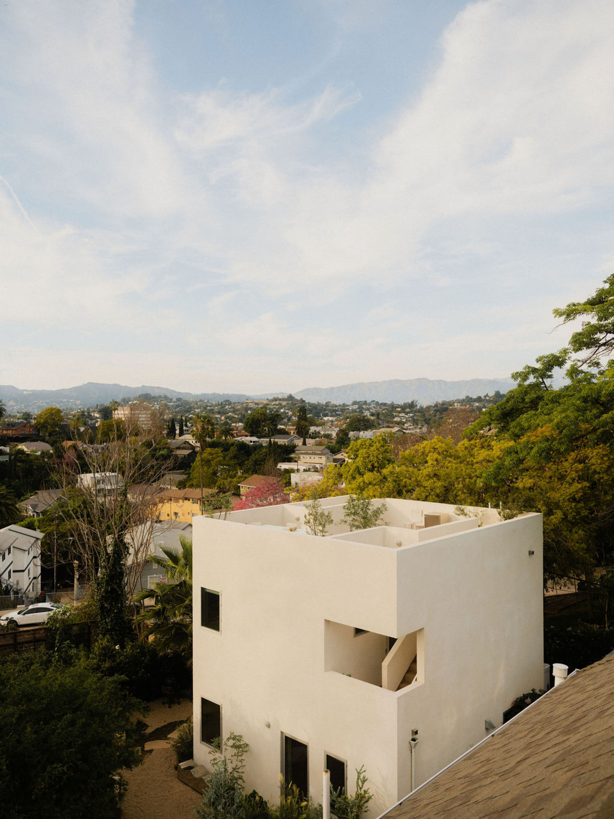 Rooftop view of a white stucco cube with a recessed stair opening and planted terrace above a hillside neighborhood under wispy clouds