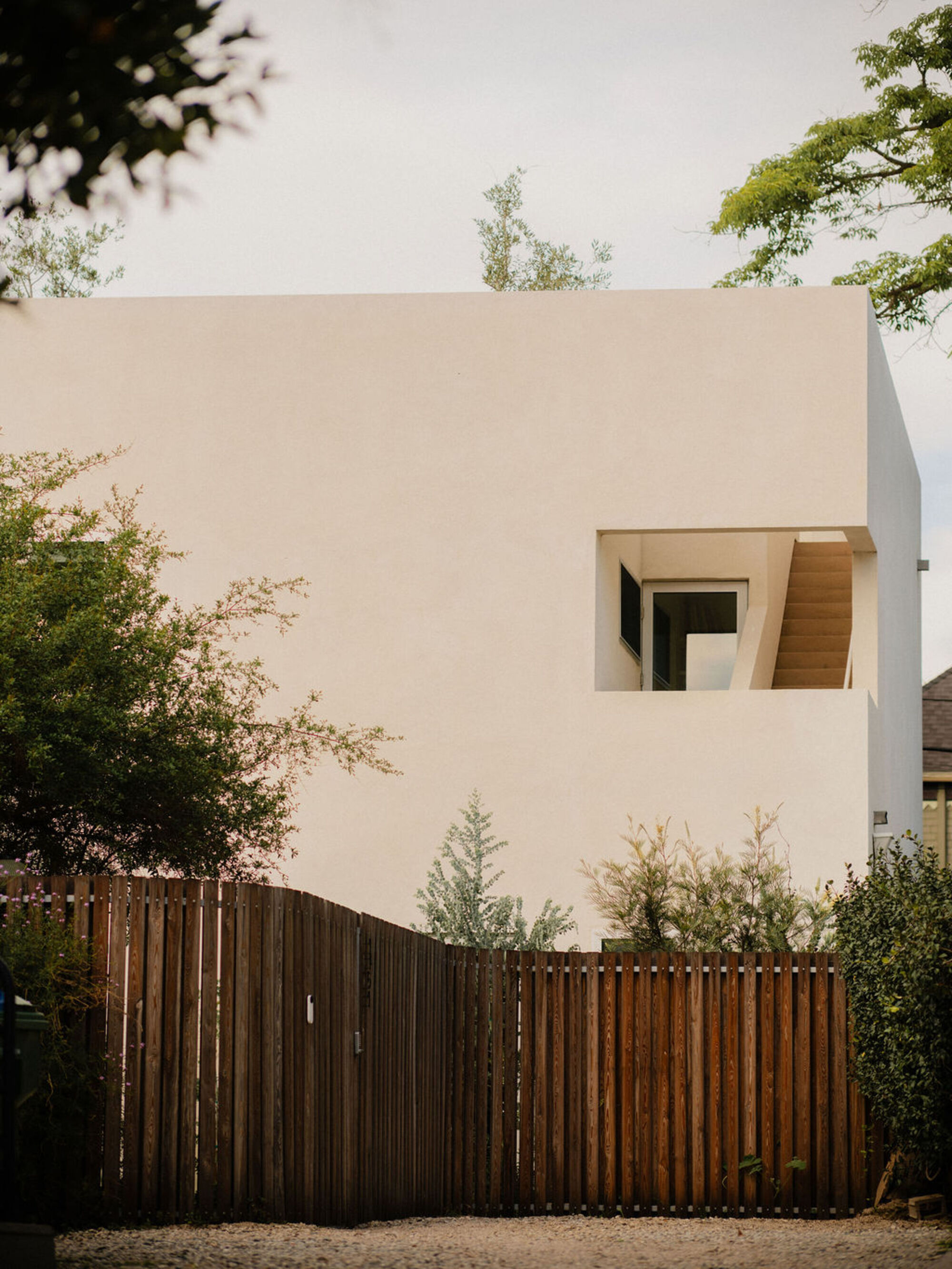 White stucco volume with a square cutout revealing a timber stair, set behind a tall slatted wood fence and gravel drive