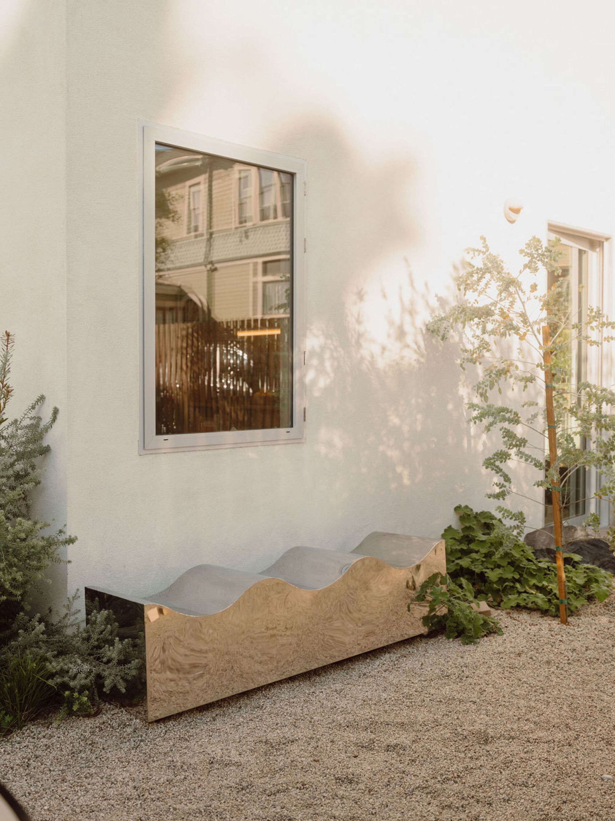 Wave-profile polished steel bench against a white stucco wall beside a framed window, with gravel ground and young trees in dappled light