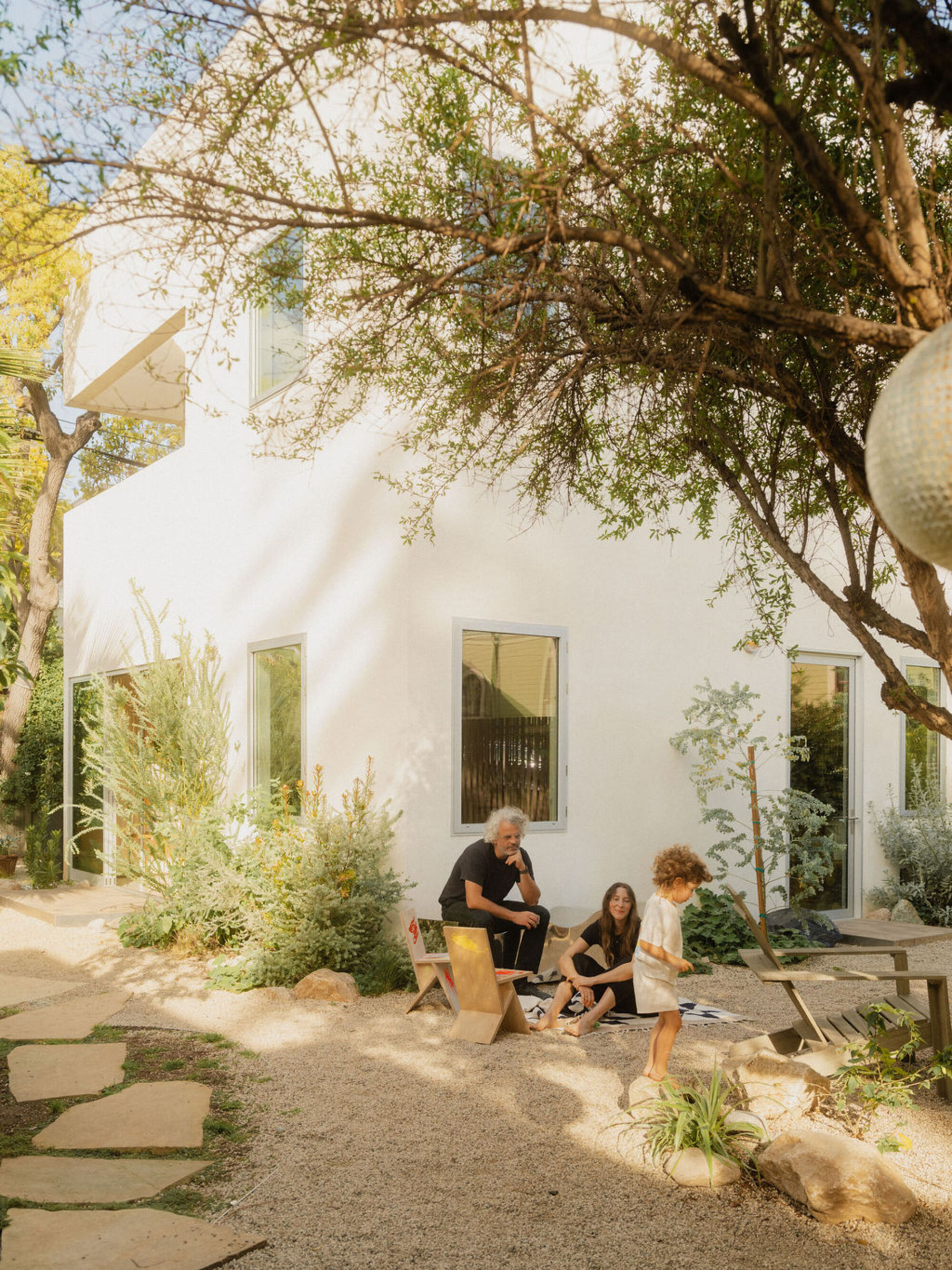 Three people seated in a gravel garden with stepping stones in front of a white house, shaded by an overhanging tree branch