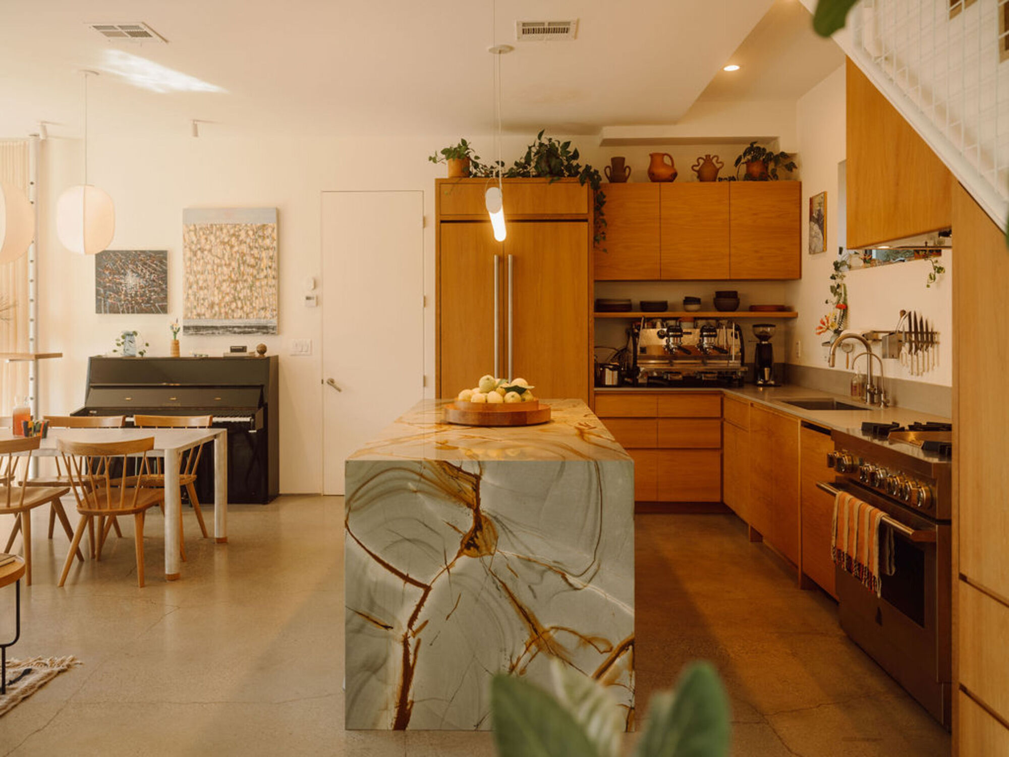 Kitchen island clad in veined green and gold stone, with timber cabinets, a espresso machine and an upright piano behind in warm light