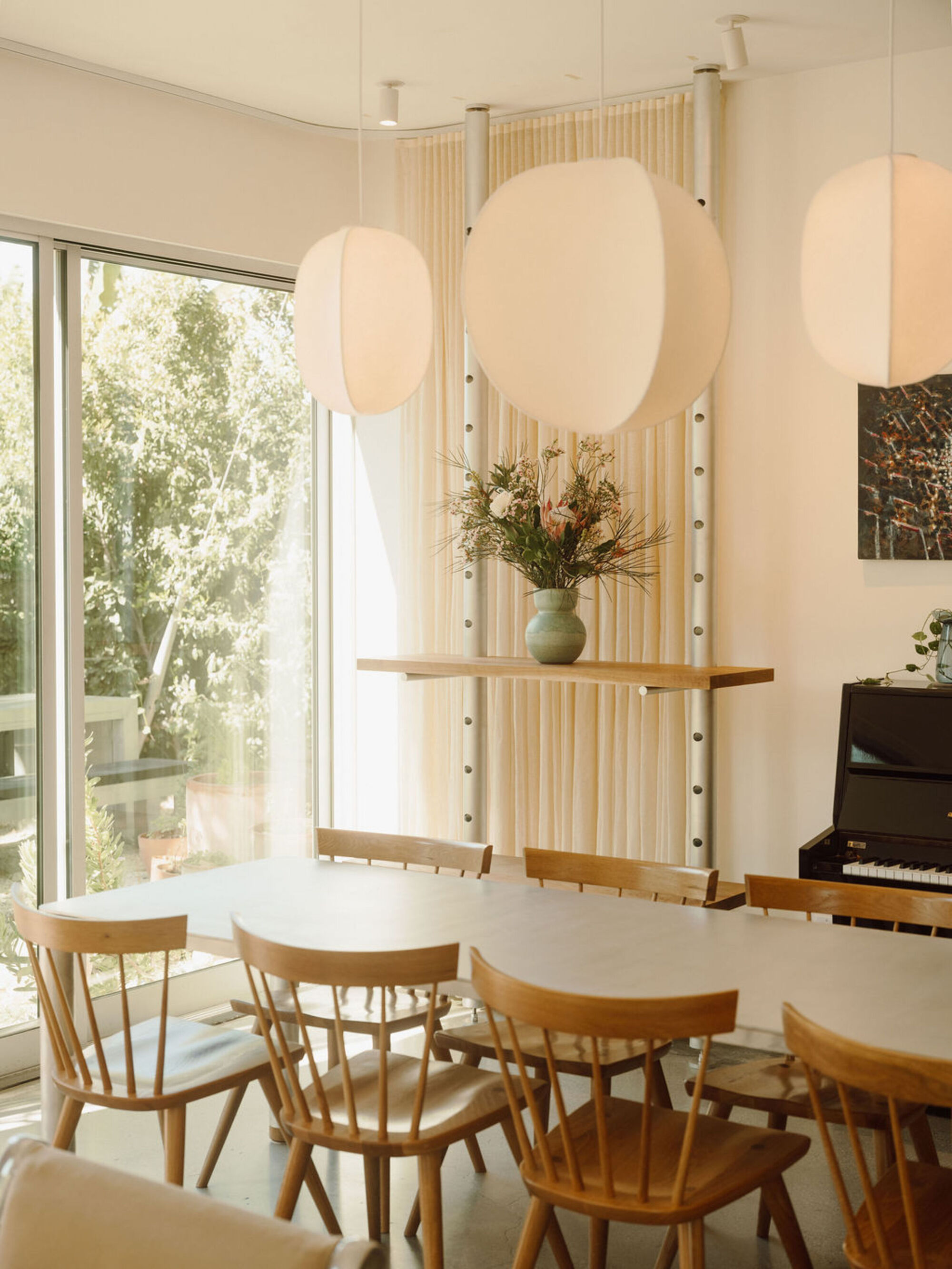 Dining table with spindle-back chairs under paper pendant lamps, beside a curtain hung between perforated steel posts and a garden glazing wall