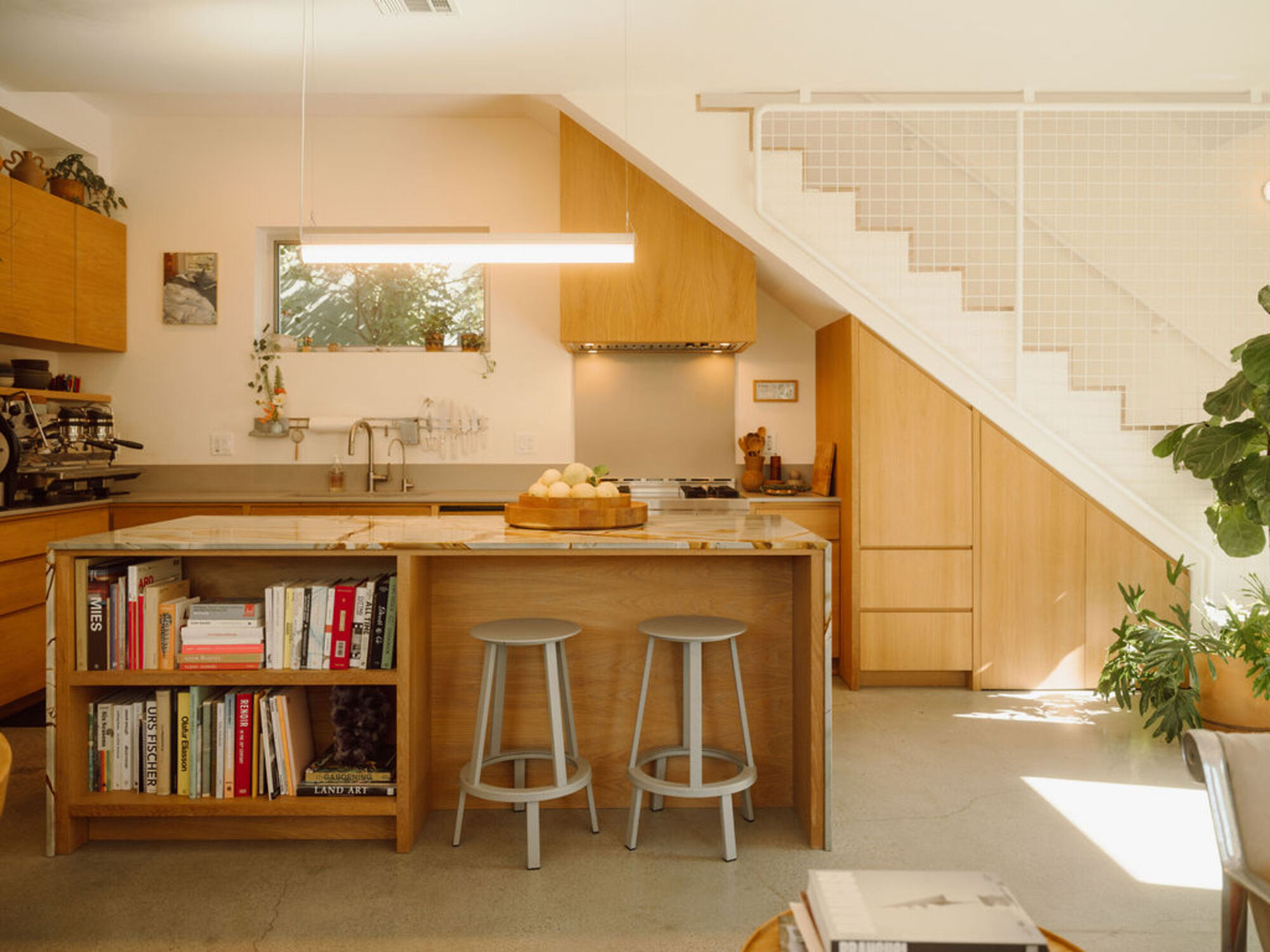 Kitchen island with a built-in bookshelf and two grey stools, beside oak cabinets tucked under the mesh-railed stair