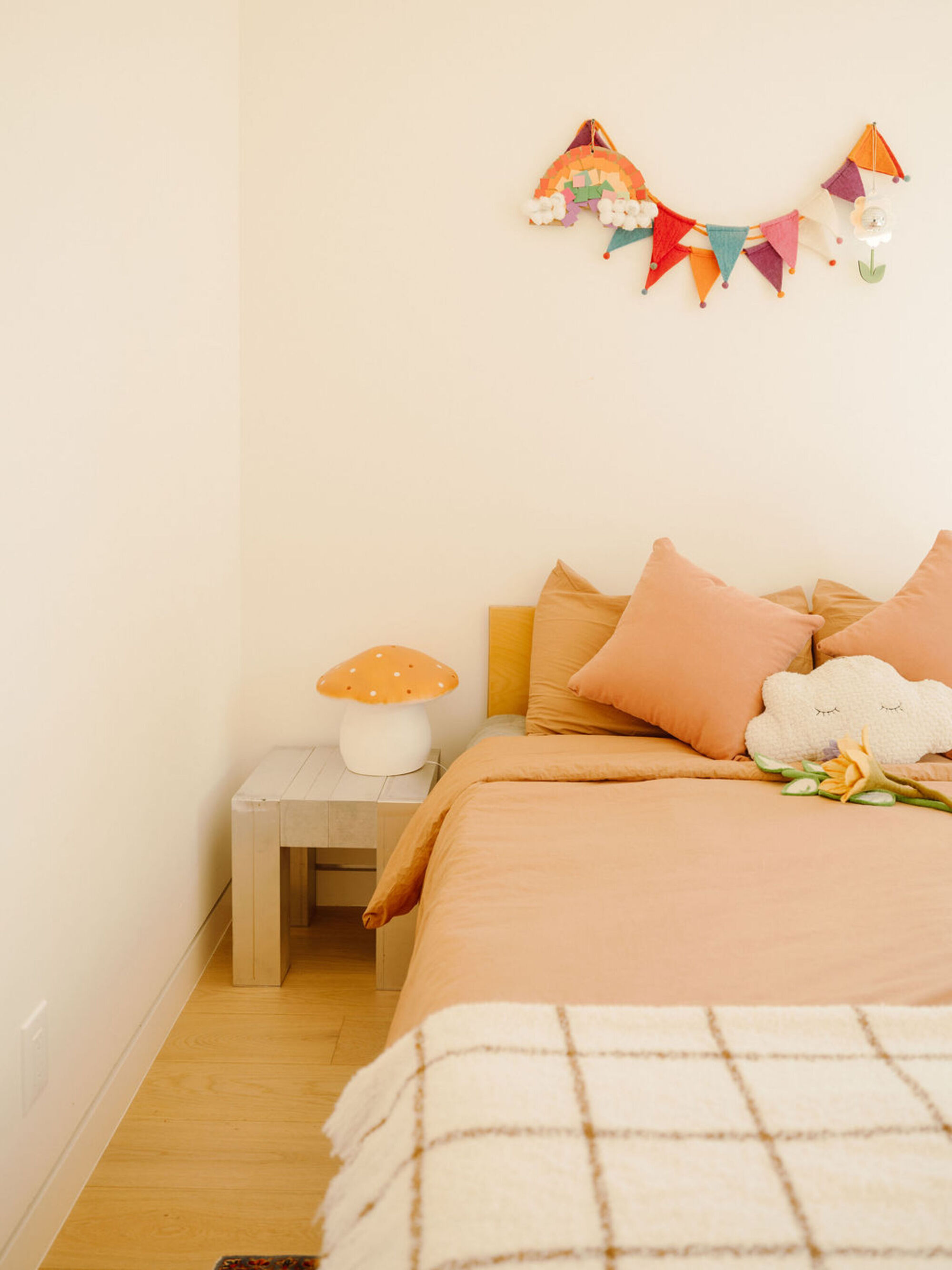 Child's bedroom corner with peach bedding, a mushroom lamp on a pale wood side table and a felt bunting on the wall
