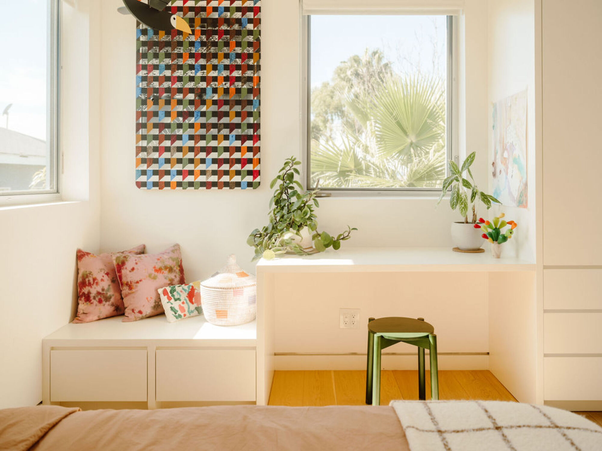 Built-in window seat and desk nook with a green stool, pink cushions and a plant in afternoon sun