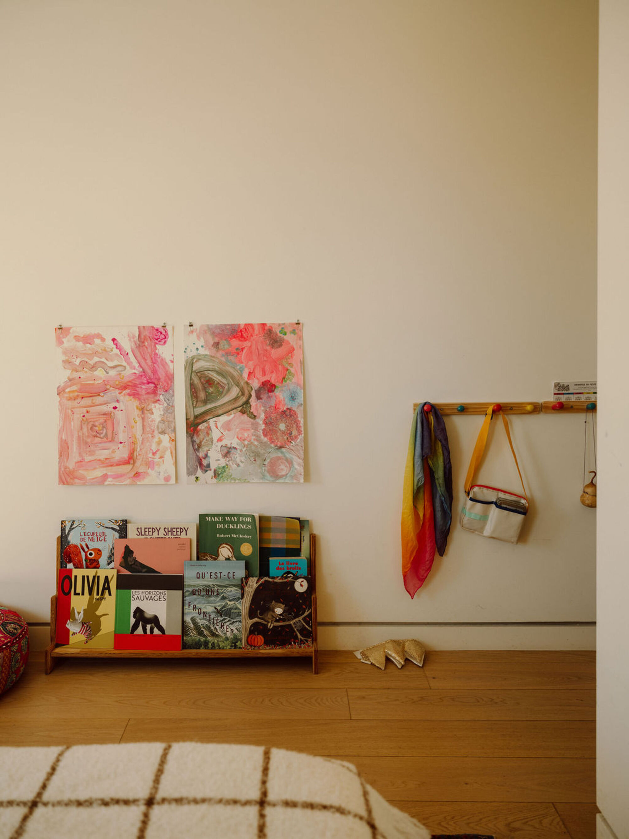 Wall with two watercolor paintings, a low front-facing book shelf and a peg rail holding a rainbow scarf and small bag