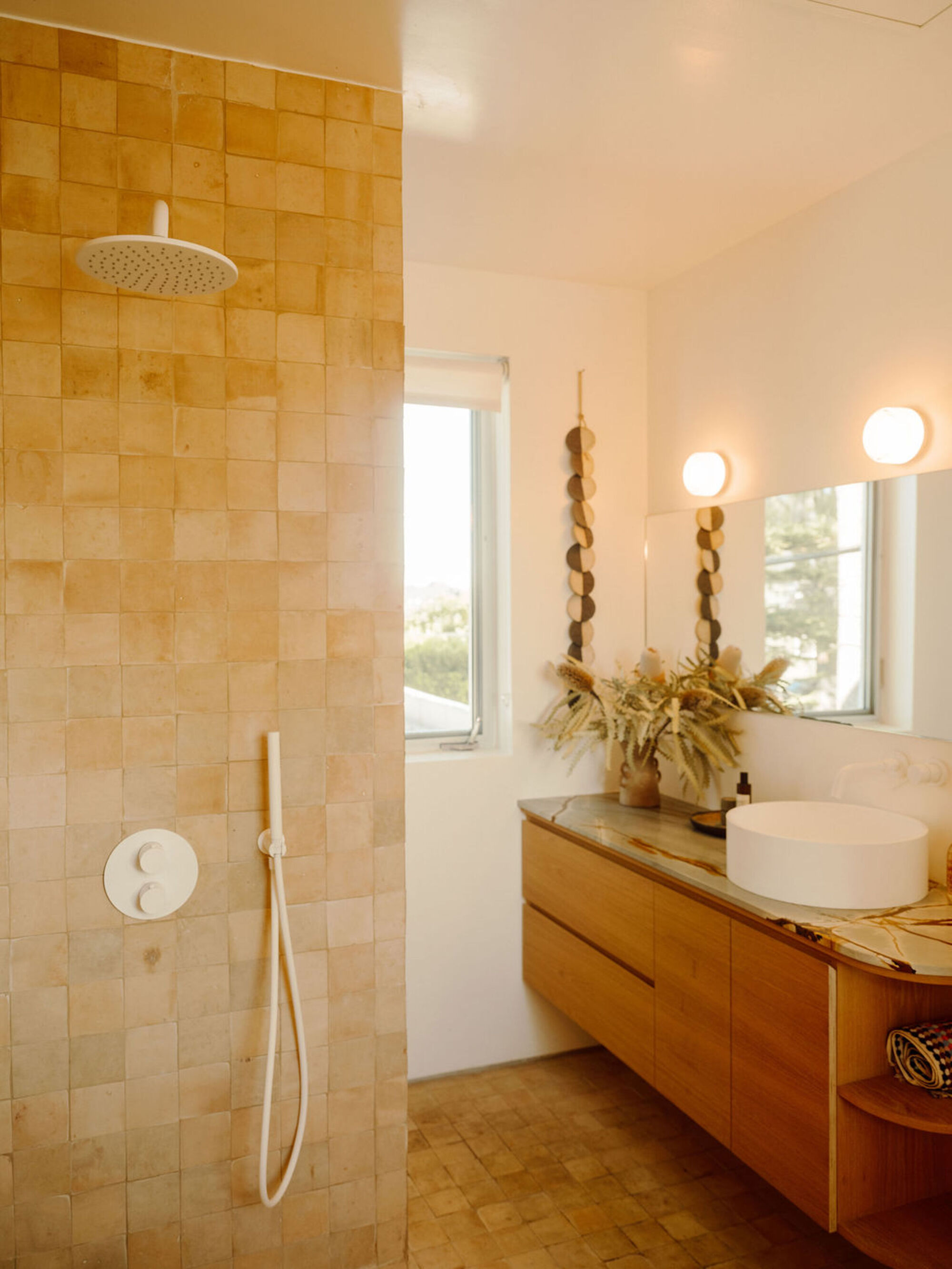 Bathroom with ochre square tile shower wall, rain showerhead and a floating oak vanity beneath a round mirror in daylight