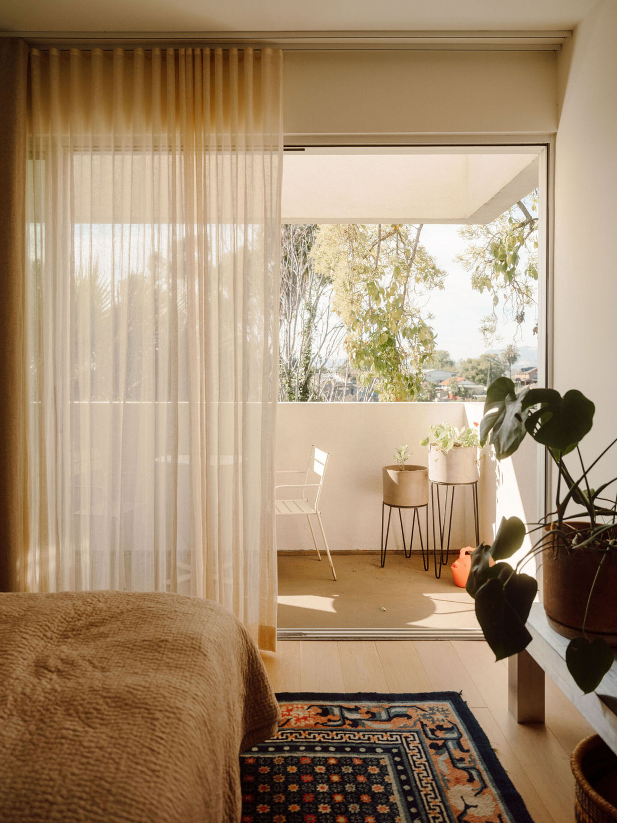 Bedroom corner with sheer curtain, patterned rug and a small balcony with potted plants in afternoon sun