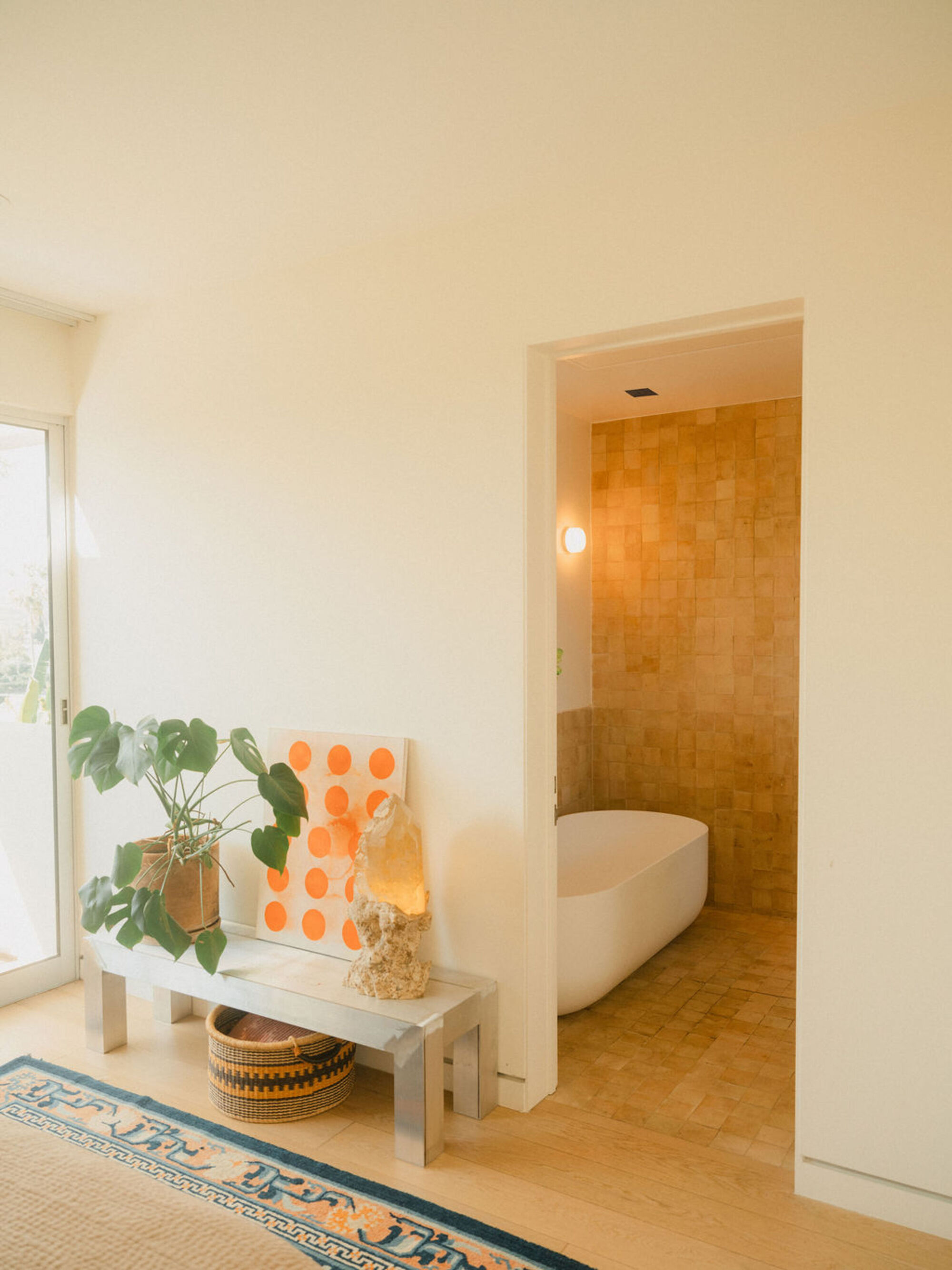 Metal bench with monstera, orange dotted artwork and a crystal lamp, opening to a bathroom with a freestanding tub and ochre tiles