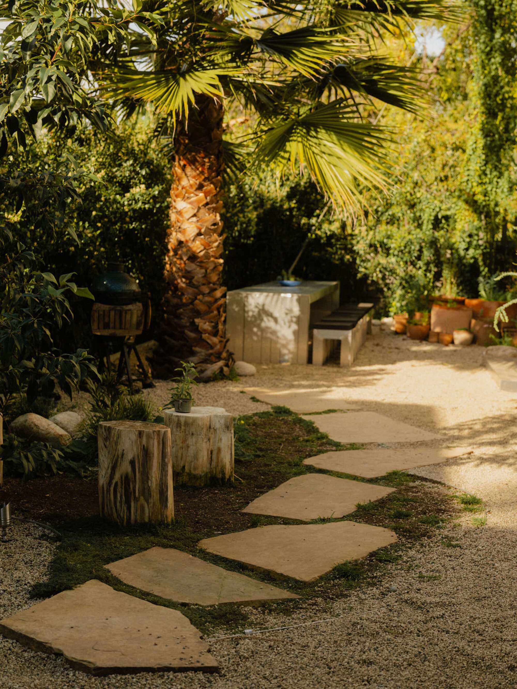 Flagstone stepping path through gravel and moss toward a concrete table and benches under a palm, in dappled light