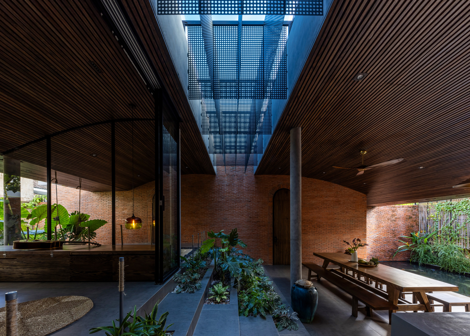 Courtyard between slatted timber ceilings with a perforated metal screen overhead, stepped planting bed and long wooden dining table in daylight