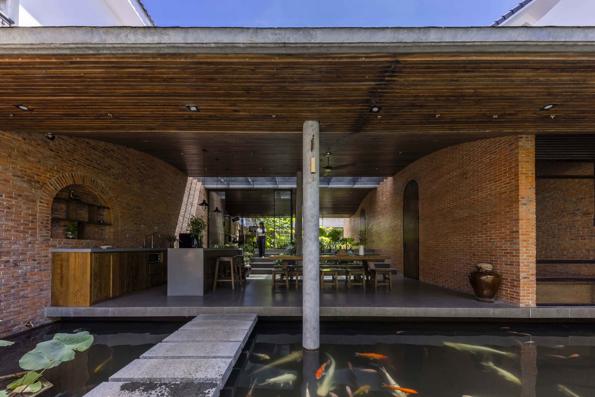 Frontal view of the open kitchen and dining space behind a koi pond, with stepping stones leading to a concrete column