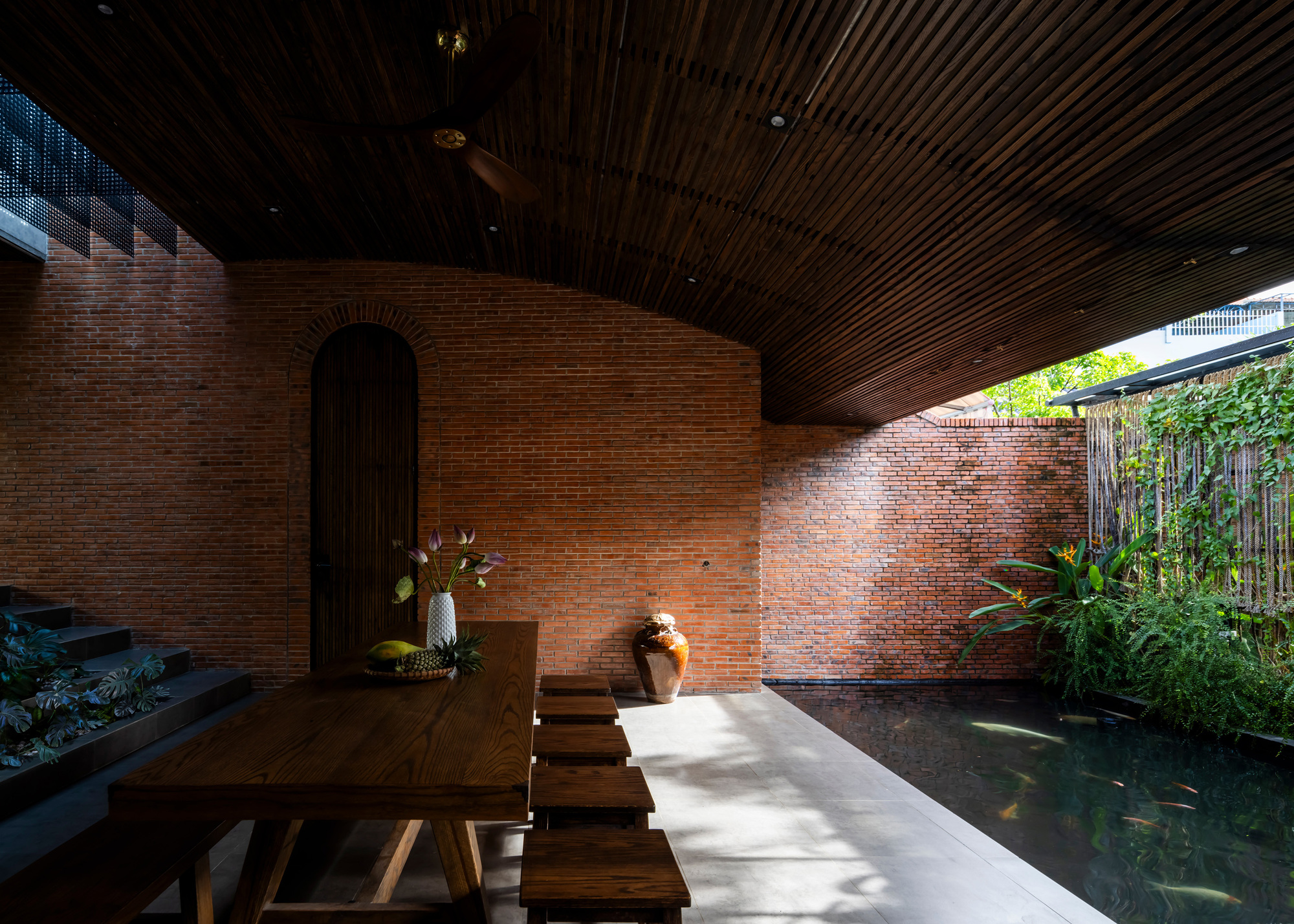 Long timber table and stools beneath a curved slatted ceiling, with dappled sunlight falling across the floor beside the koi pond
