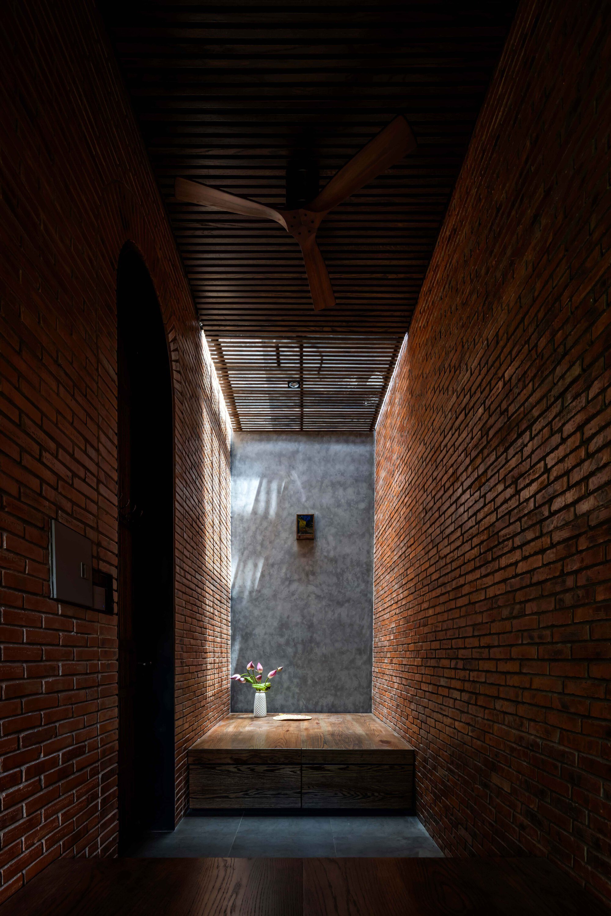 Narrow brick passage ending in a timber bench and vase of flowers, with a ceiling fan and light spilling from above