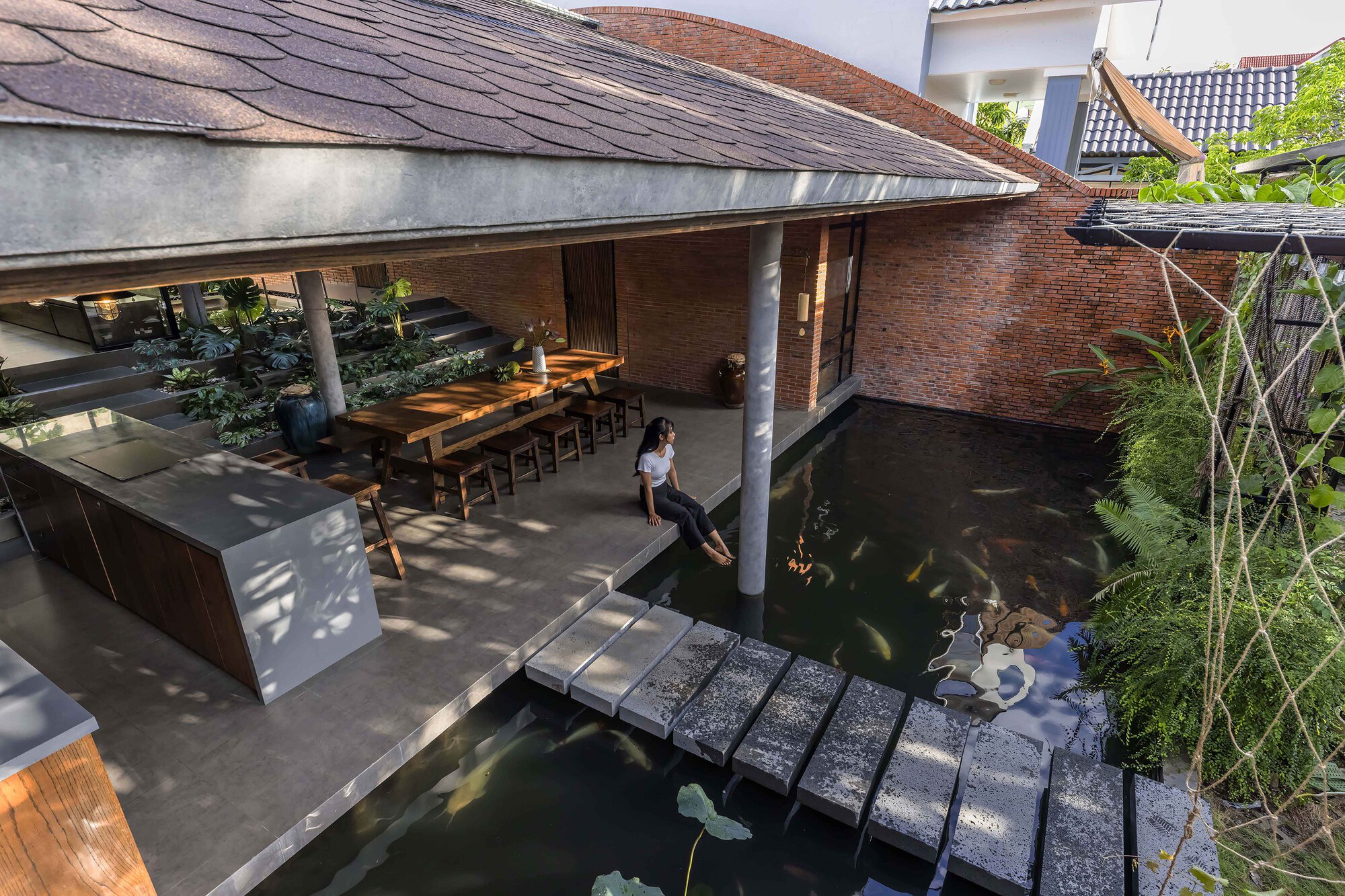 High view of a woman seated at the pond edge beside stepping stones, with the open kitchen and dining table behind