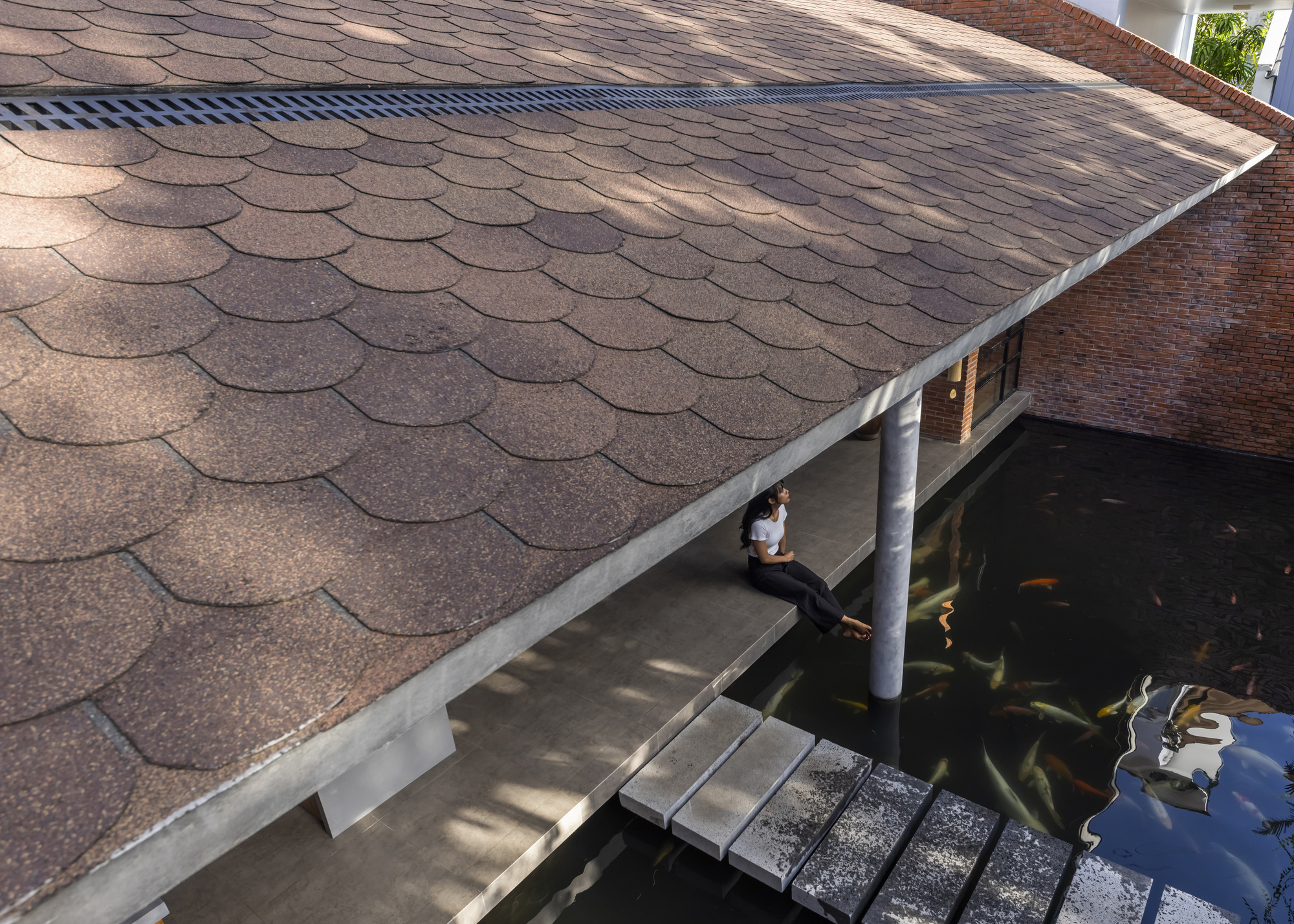 Close view of the shingled roof and its drainage strip, with a woman sitting under the eave above koi swimming in the pond