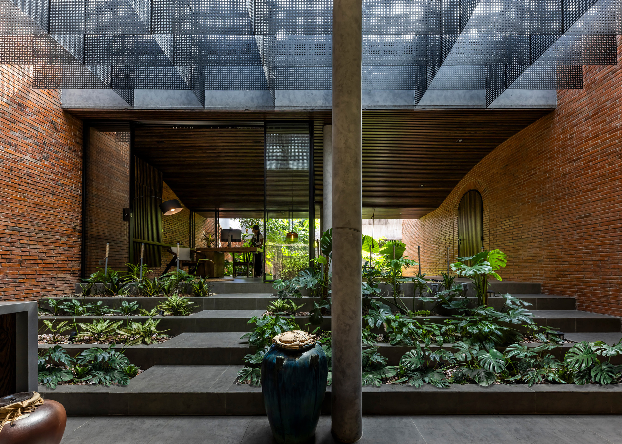 Stepped planter terraces of monstera rising toward a glazed workspace, flanked by curved brick walls and a perforated metal canopy overhead