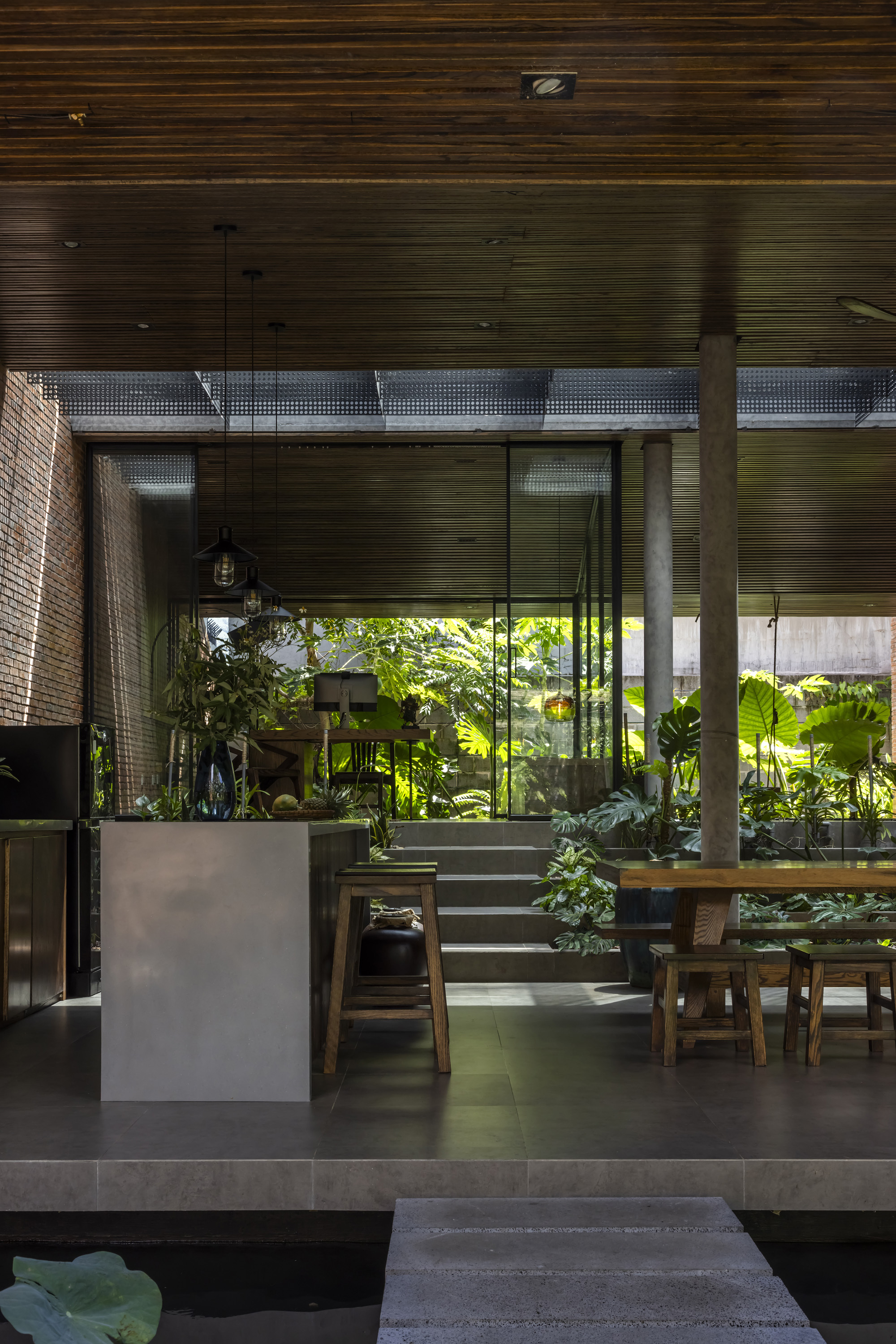 Open dining pavilion with timber slat ceiling, concrete column and stone stepping path over a pond, with green light filtering in