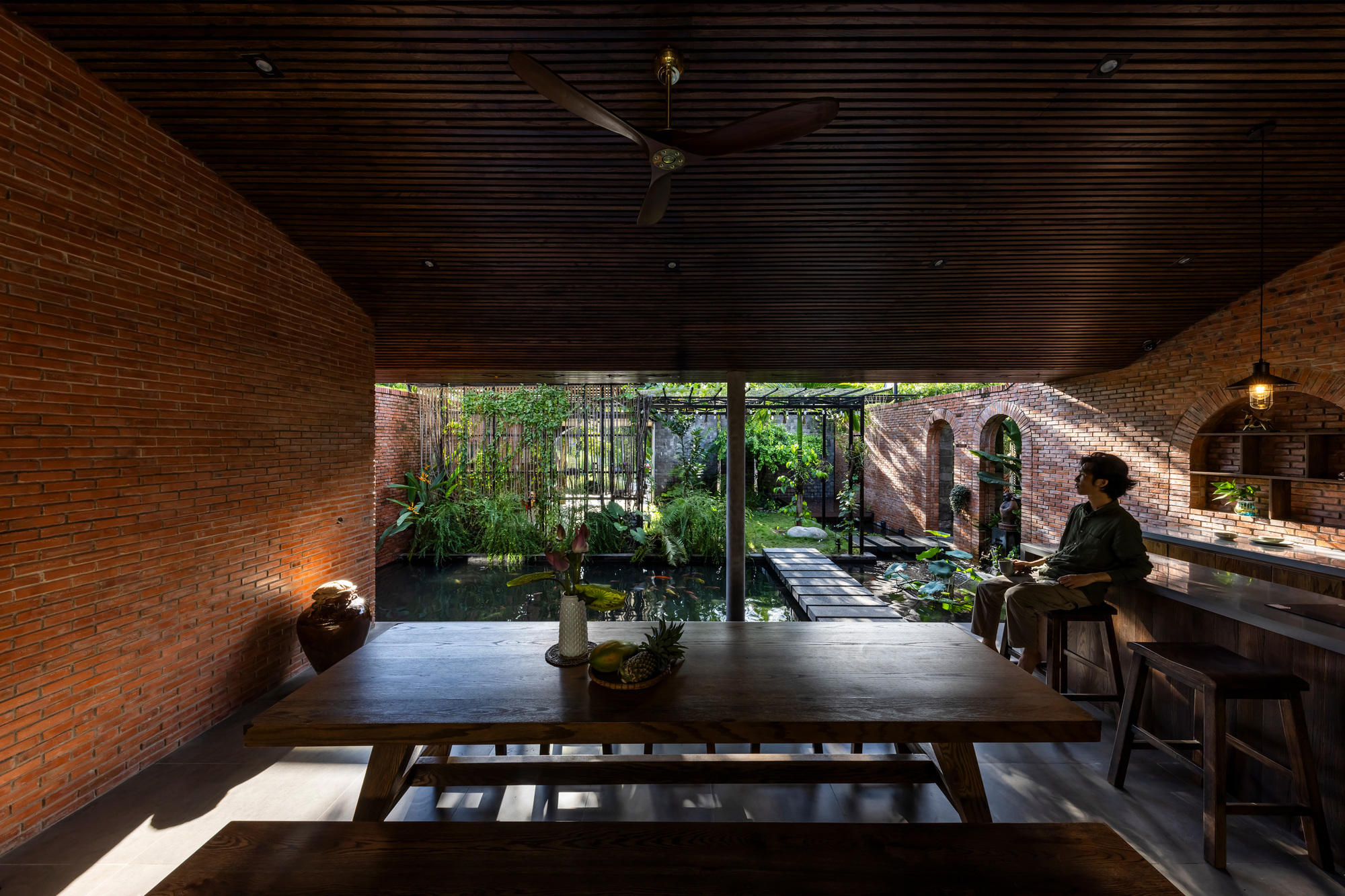 Long timber table under a slatted ceiling facing a lily pond, with a seated man at the brick-arched bar in afternoon light