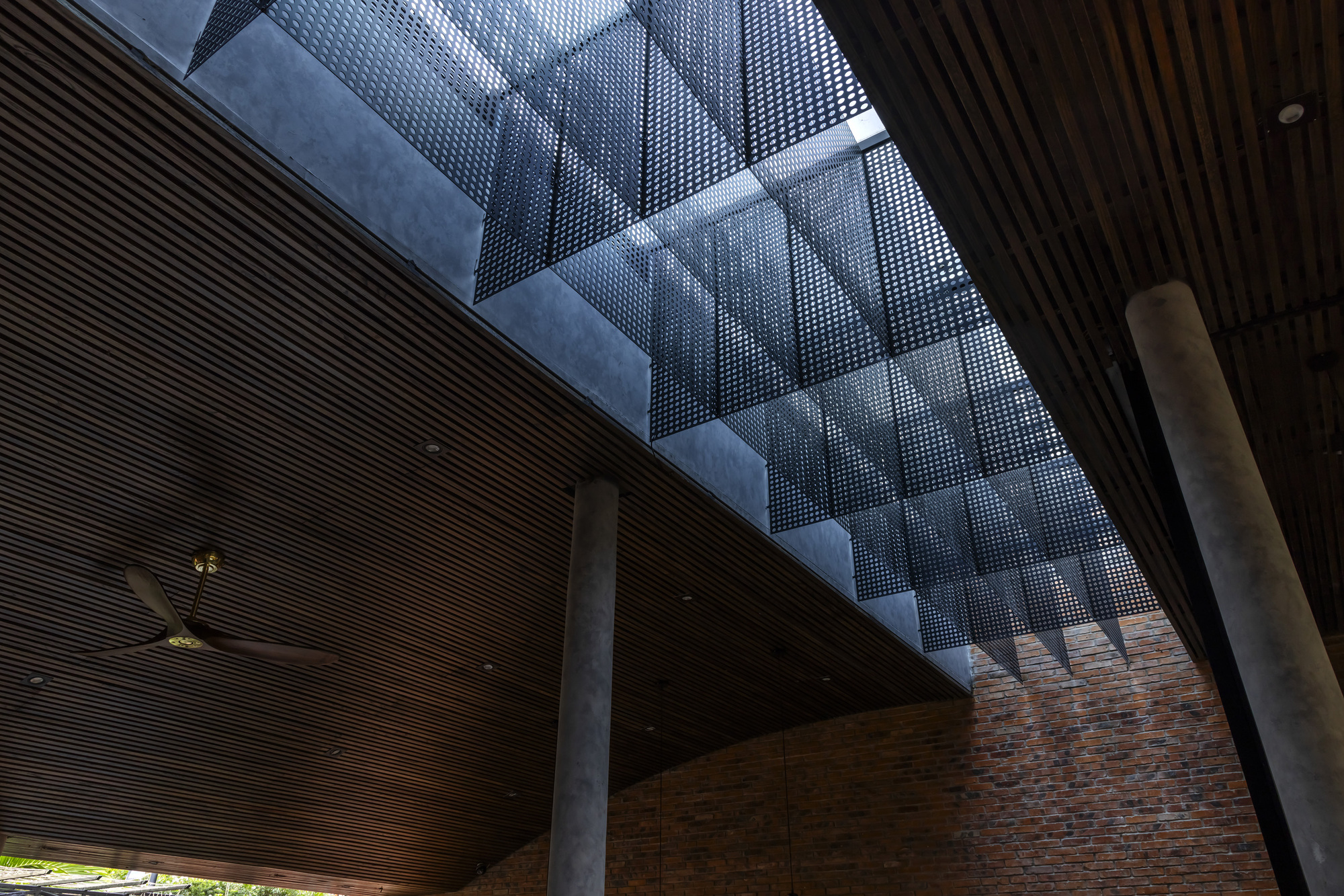Upward view of a perforated metal screen skylight above timber slat ceilings, concrete columns and a brick wall in dappled daylight