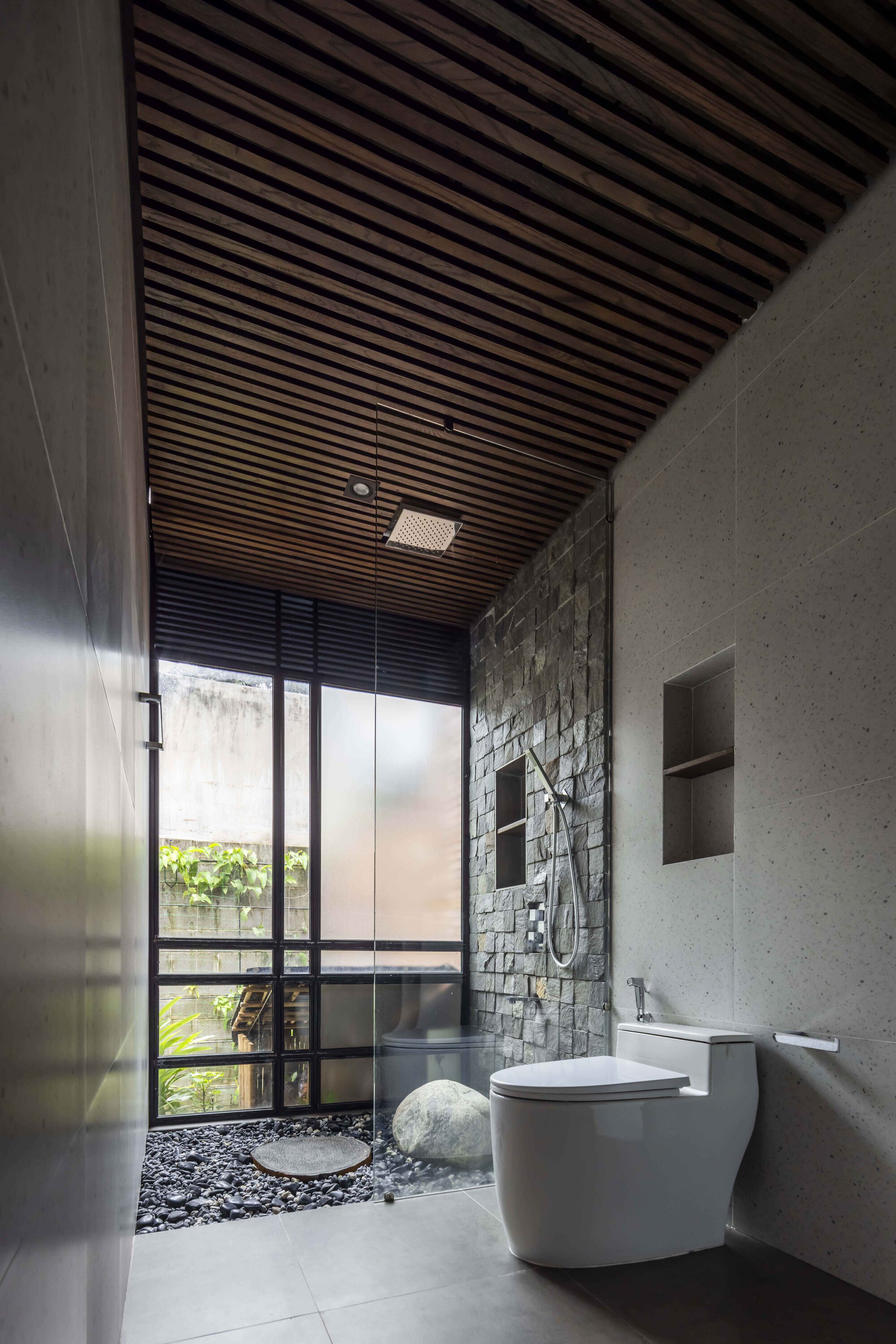 Narrow bathroom with a glass-enclosed pebble shower, stone-clad wall and slatted timber ceiling, with garden greenery visible through the window