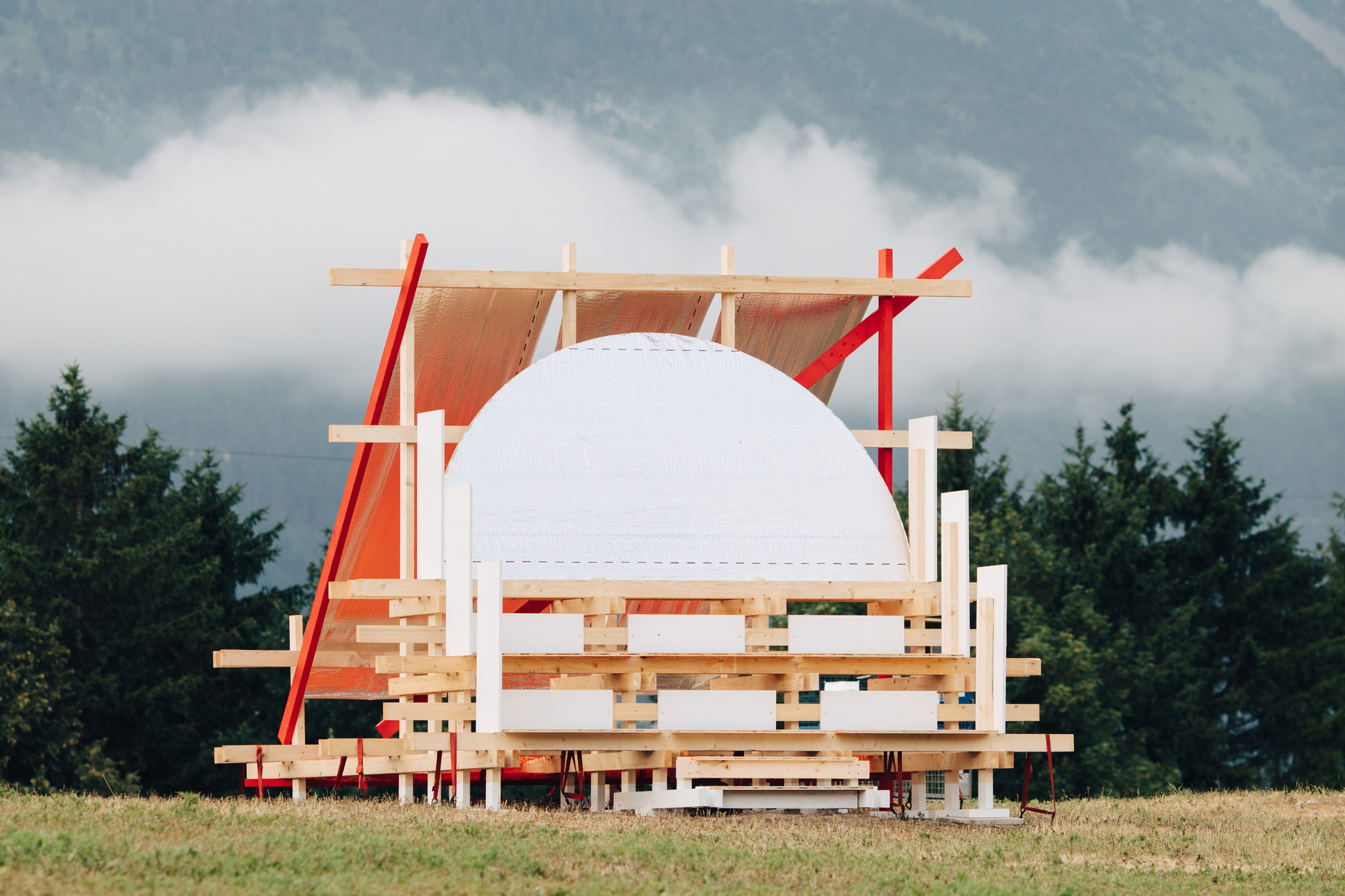 Stacked timber pavilion with a white domed form and red diagonal braces, set against low cloud and forested slopes