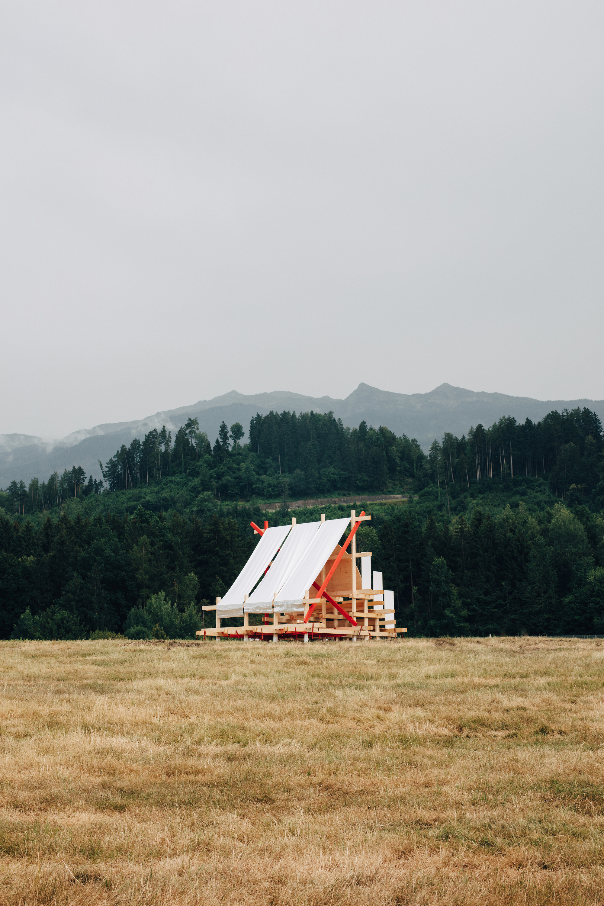 Distant timber pavilion with white fabric roof panels and red braces in a dry hay meadow under overcast sky