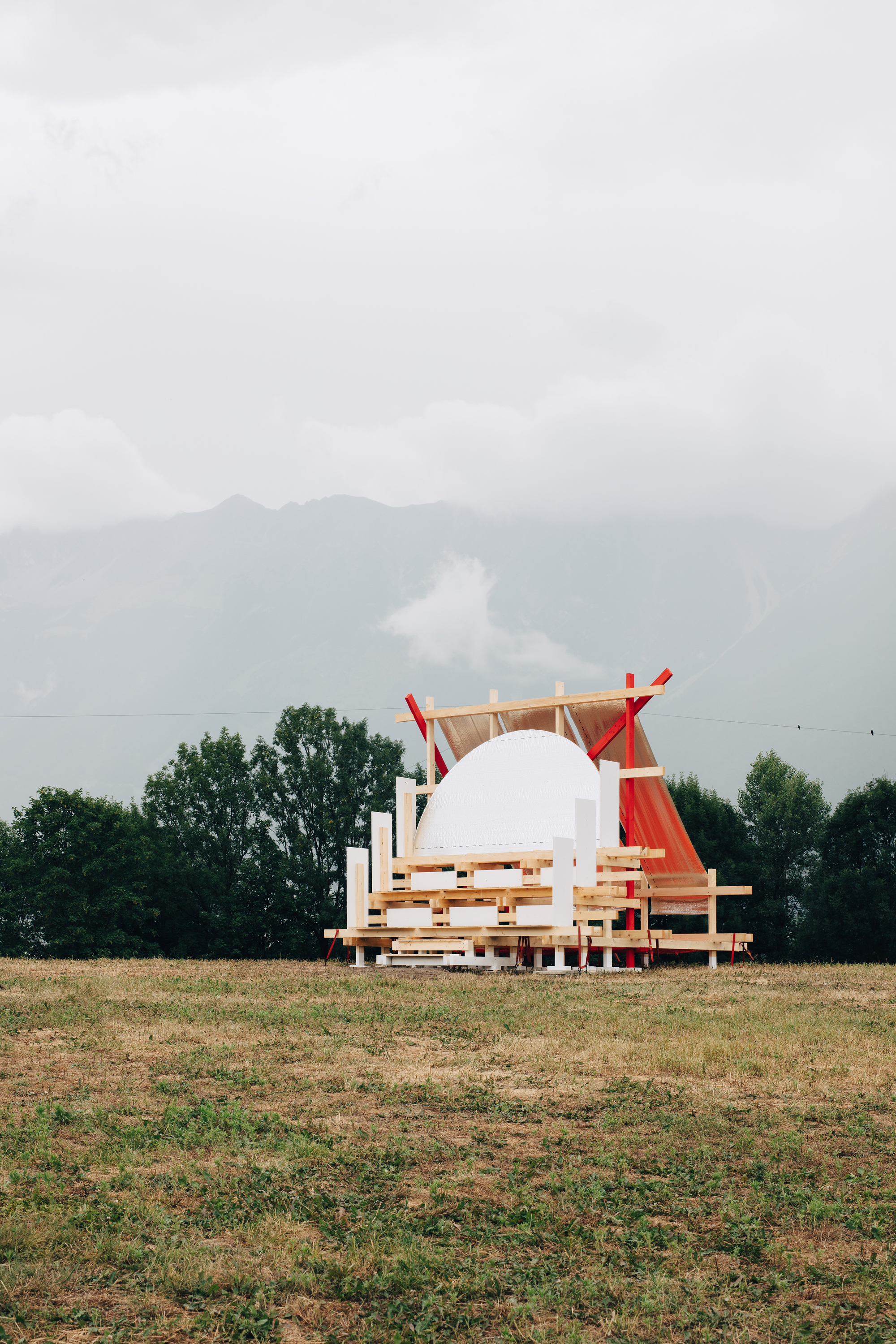 Layered timber frame with a white half-dome and orange fabric screen, seen across a meadow in drifting mist