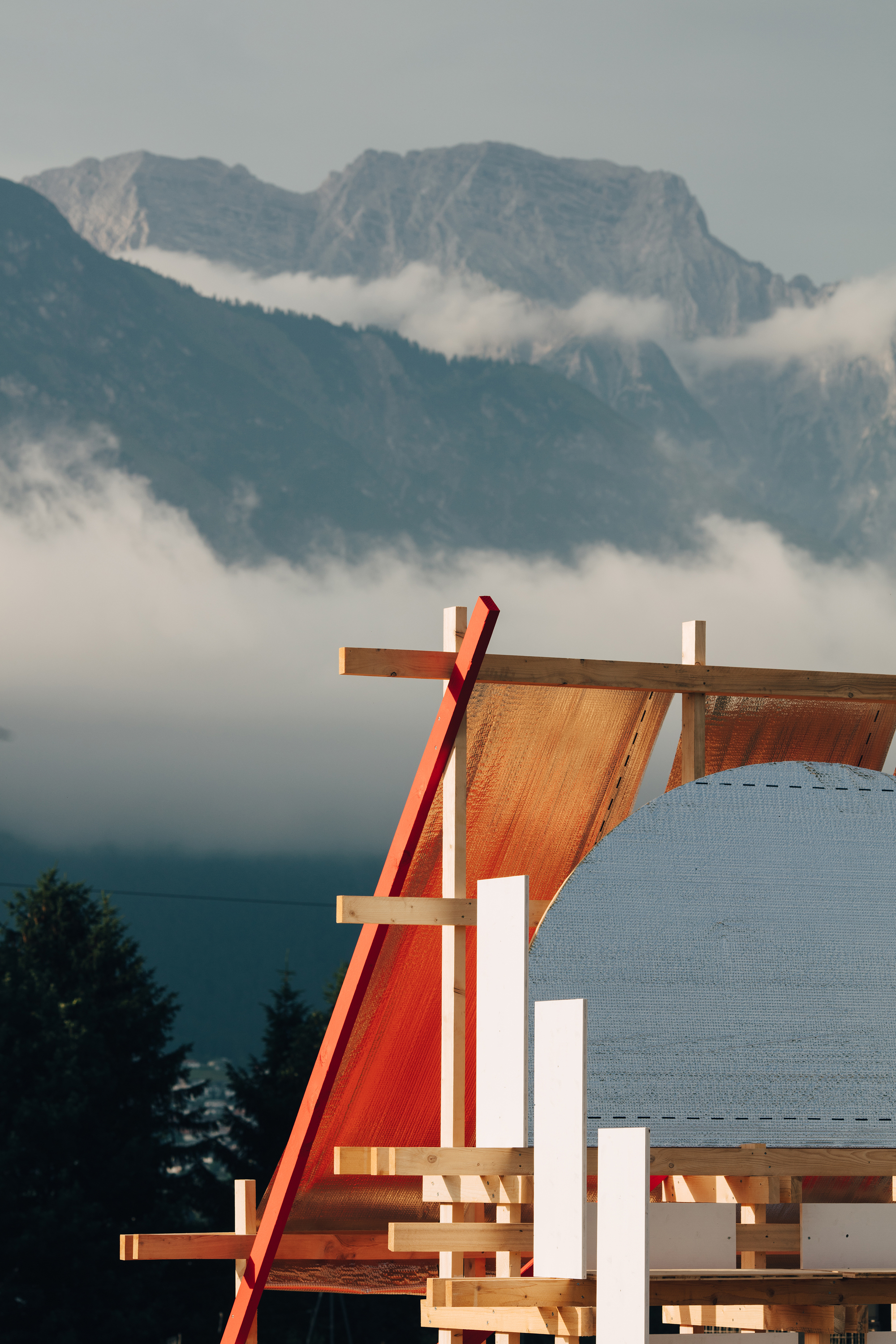 Timber frame with a red diagonal brace and orange membrane roof, set against mountains and low drifting cloud