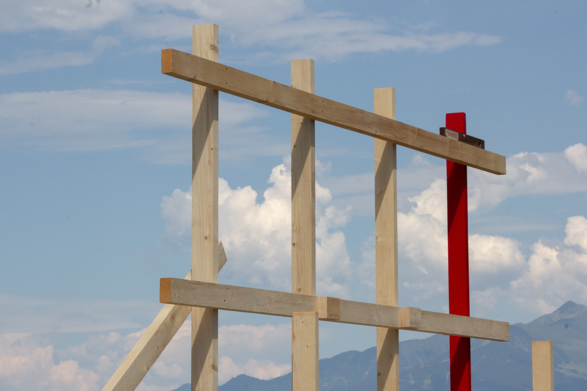 Vertical timber posts and horizontal beams beside a red post, framed against blue sky and distant mountains