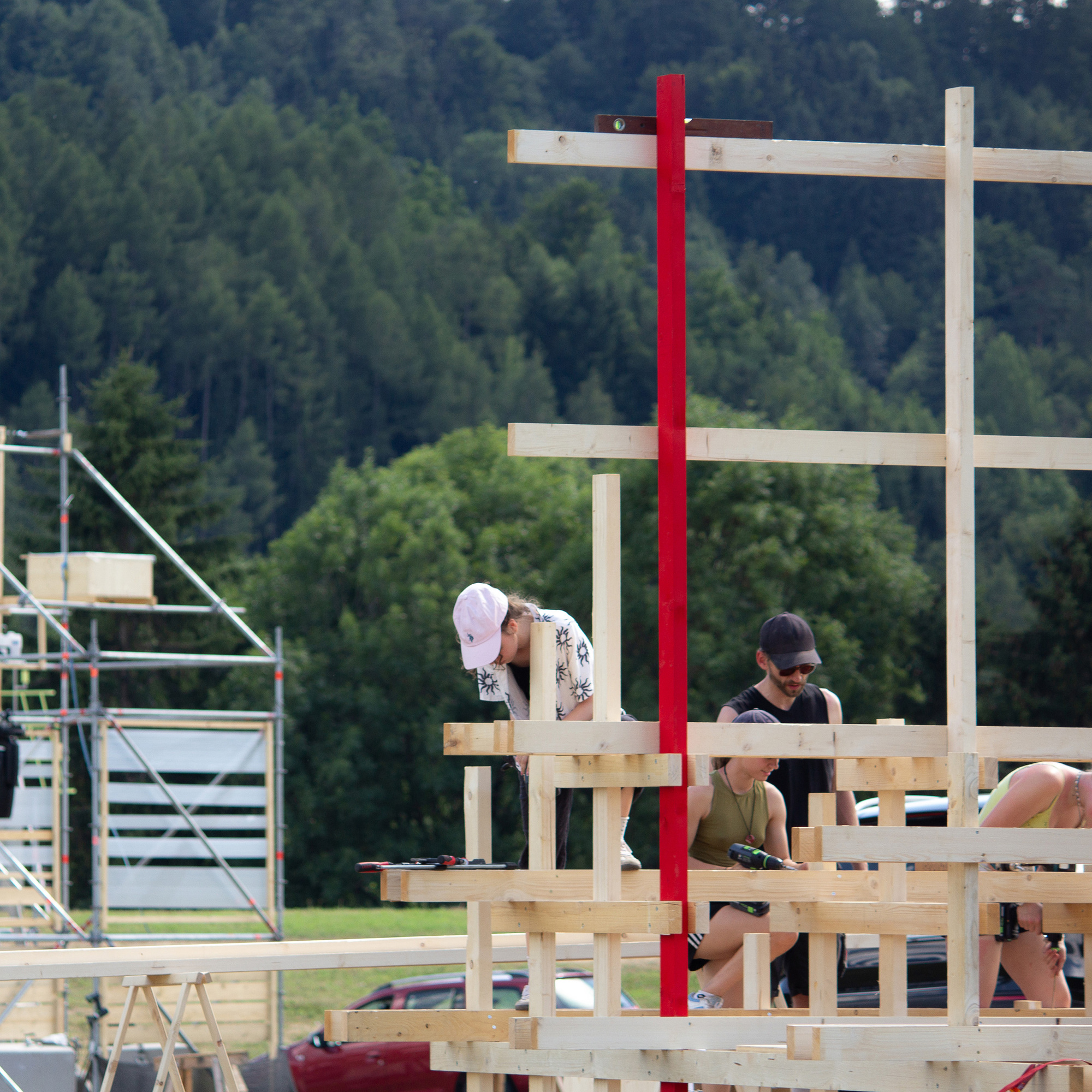 Builders working on a timber scaffold-like frame with a red post, in front of a forested hillside