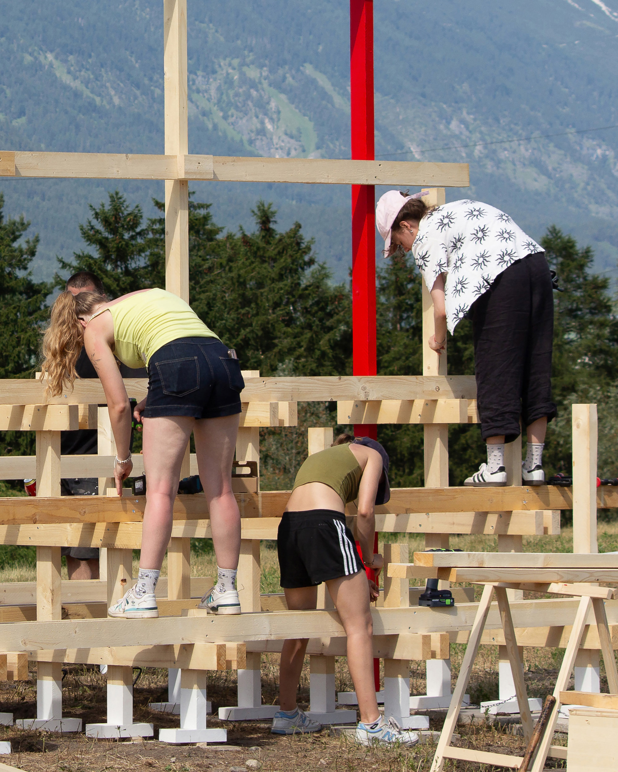 Three people fastening timber beams on a low frame with white footings, on a sunny day below forested slopes