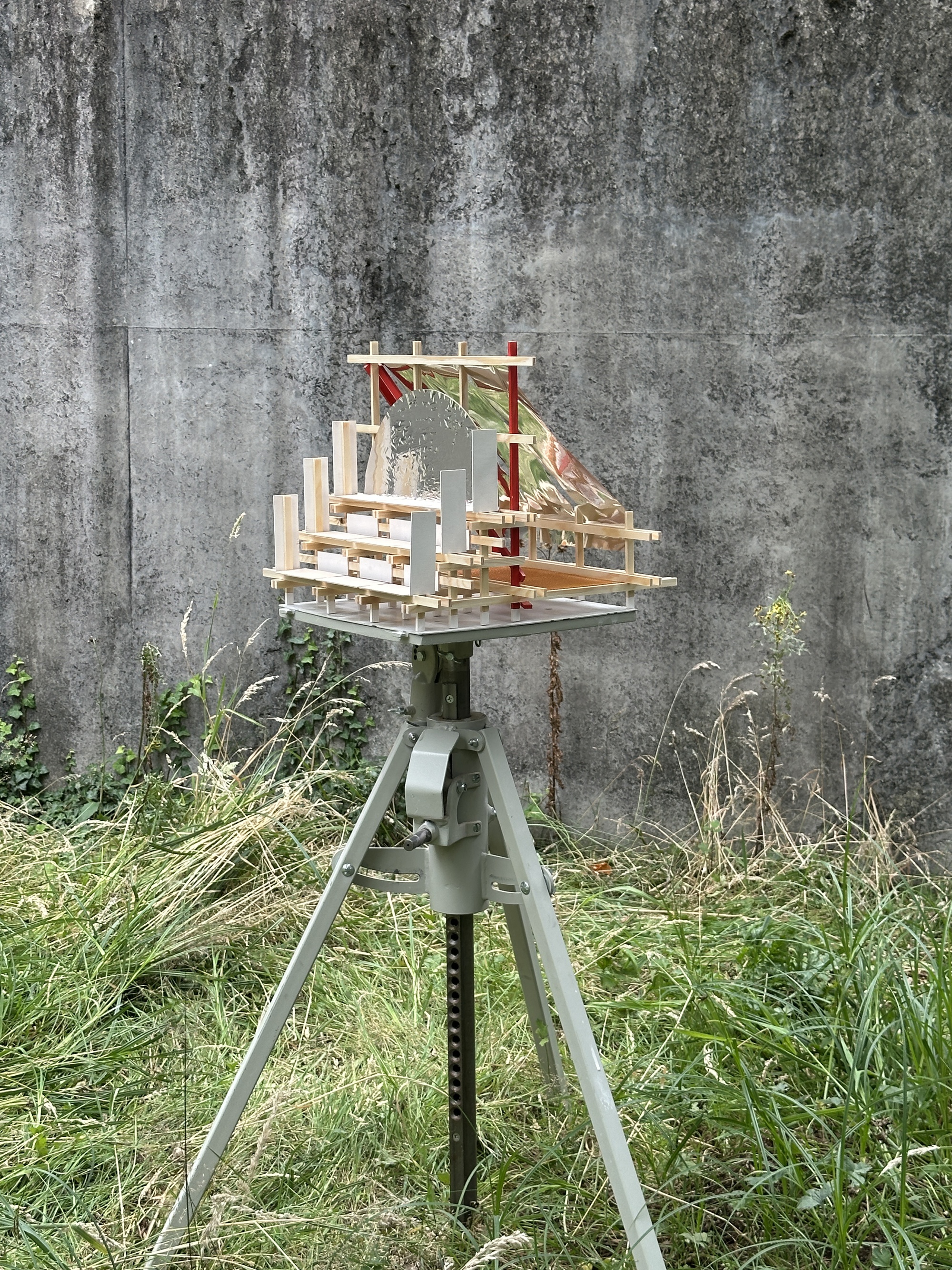 Small timber and mirrored-sheet model mounted on a metal tripod in tall grass before a weathered concrete wall