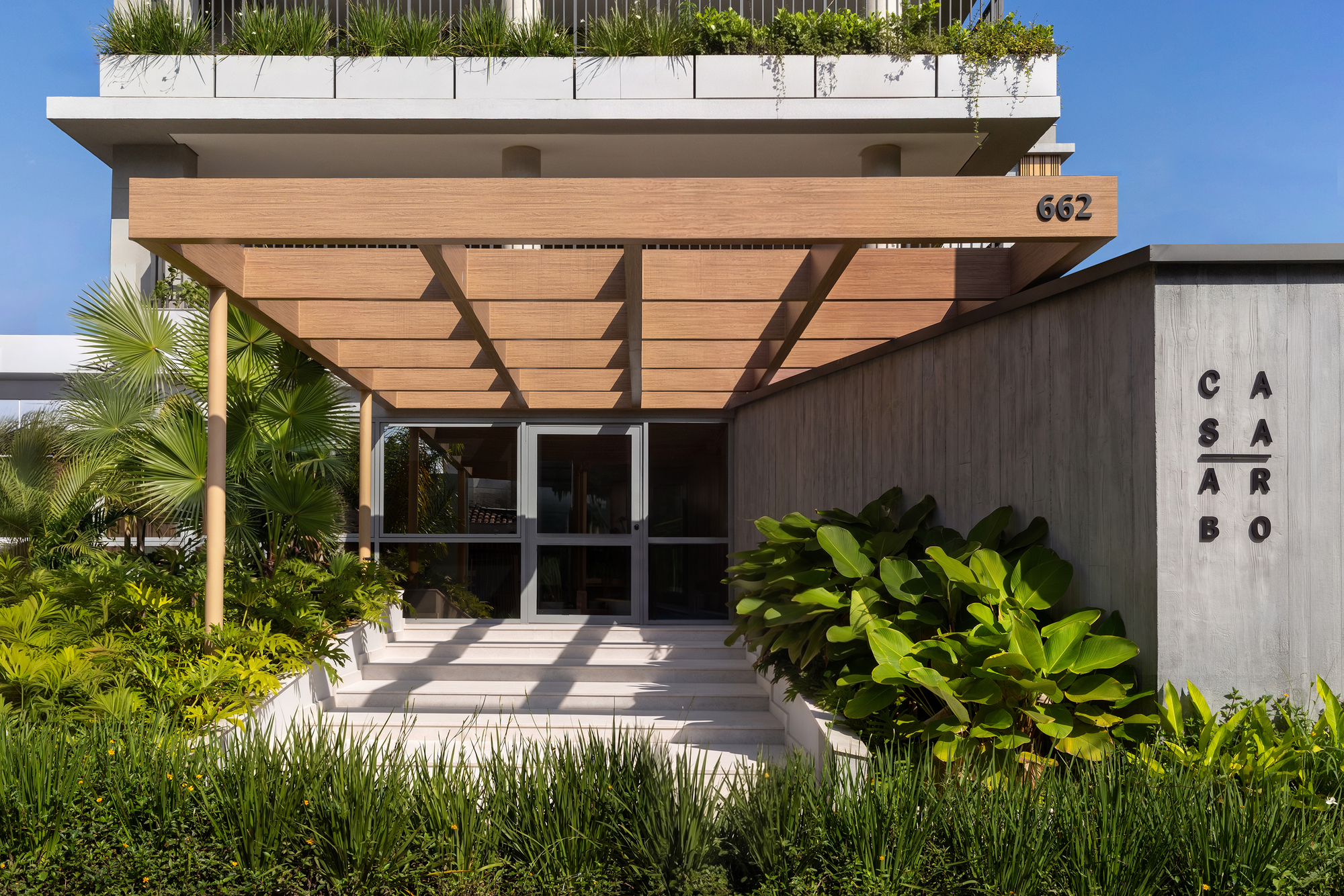 Entrance canopy of timber slats over glass doors and steps, flanked by a concrete wall and broad-leaved plants in bright sun