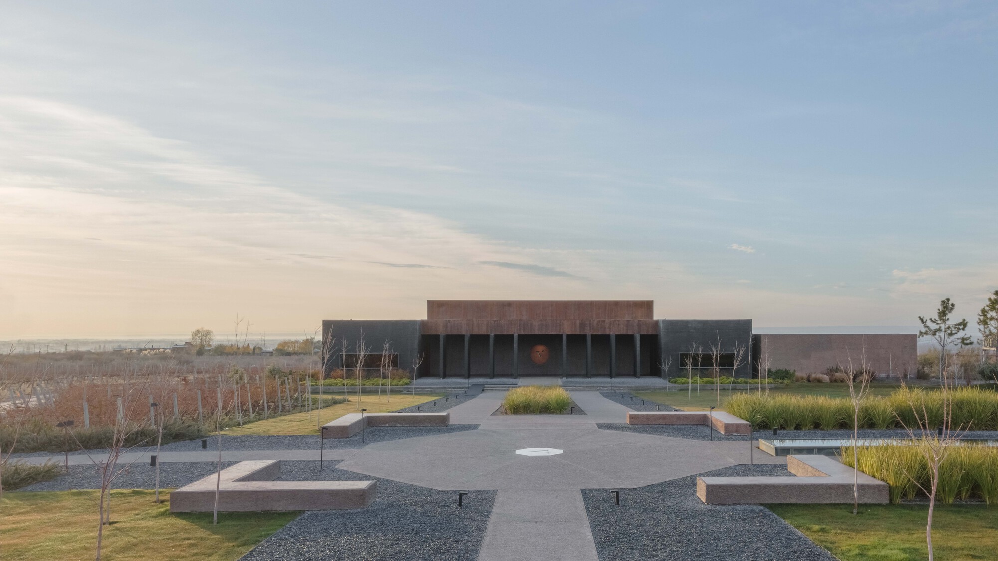 Symmetrical approach to a low weathering-steel and concrete pavilion across a gravel court with stone benches, grasses and soft dusk light