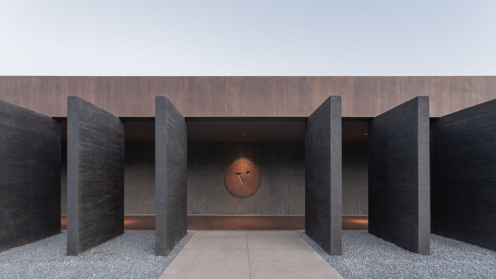 Entrance framed by tall board-formed concrete slabs under a rusted steel band, with a circular emblem on the rear wall