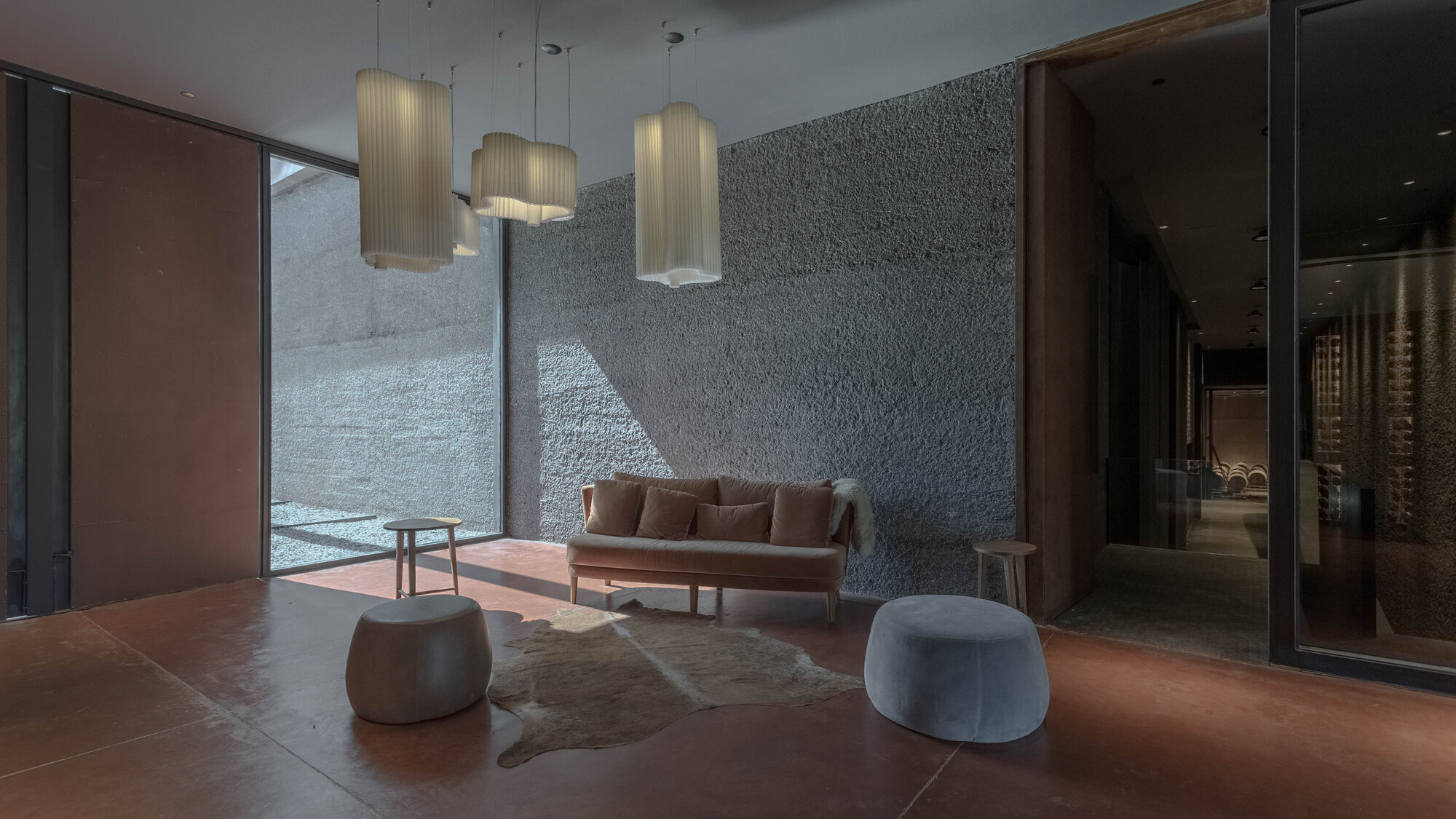 Lounge with a velvet sofa, round pouf seats and pleated pendant lamps against a textured grey wall, light falling from a side courtyard