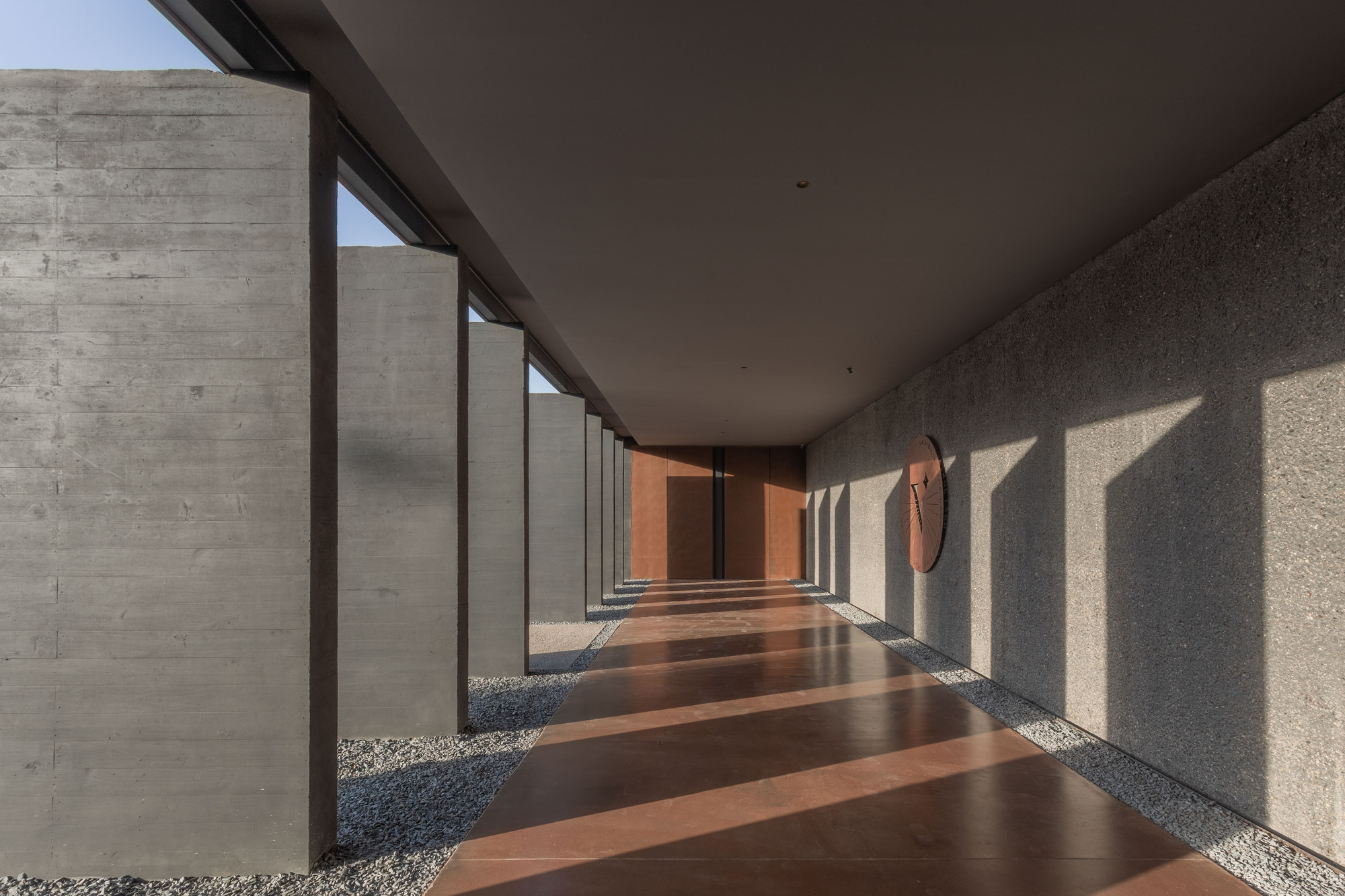 Covered walkway lined with concrete piers and a red-brown floor, late sun casting long shadows along the textured wall